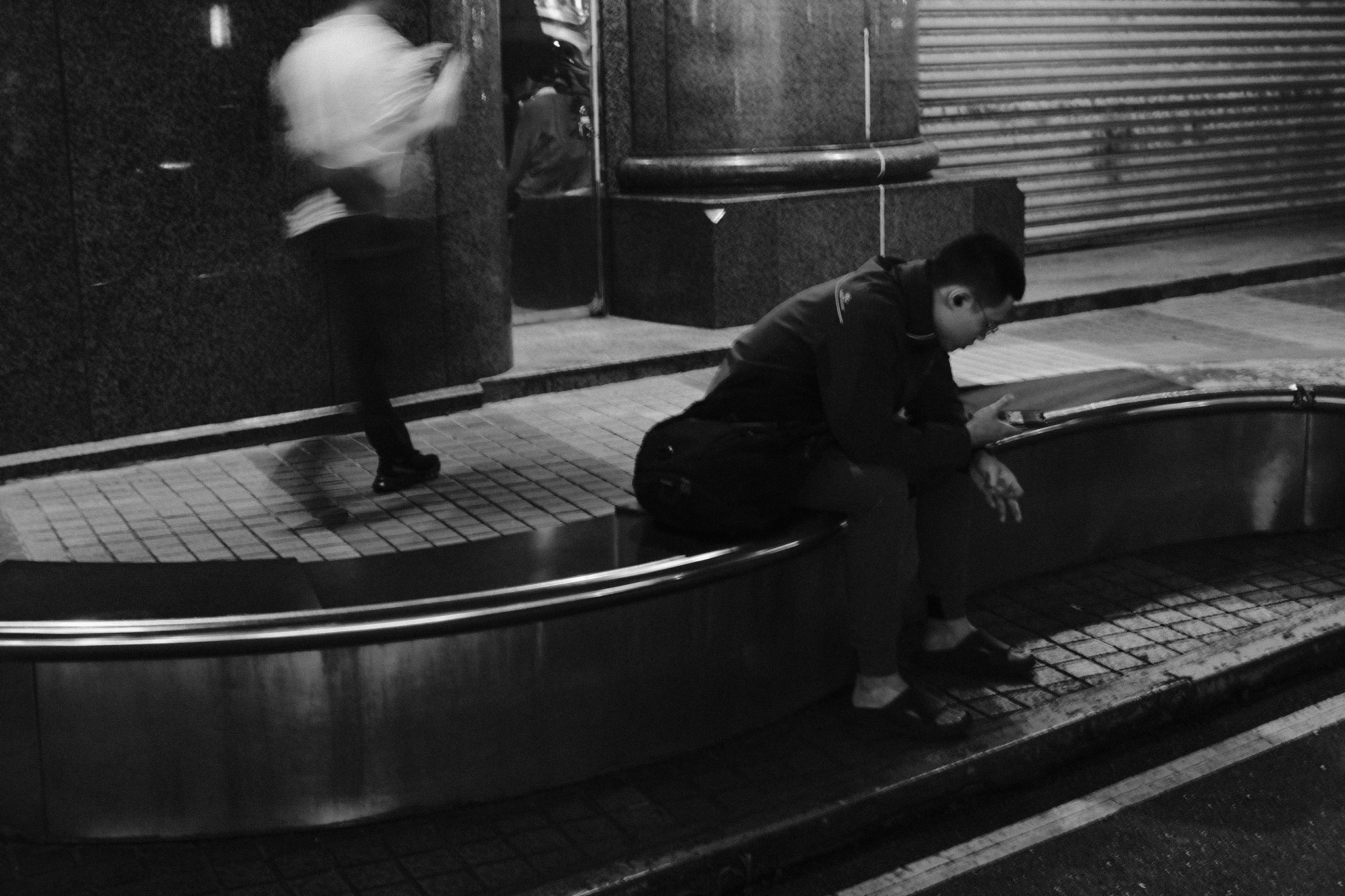 Night street motion blur with a seated figure and passing pedestrian in Taiwan