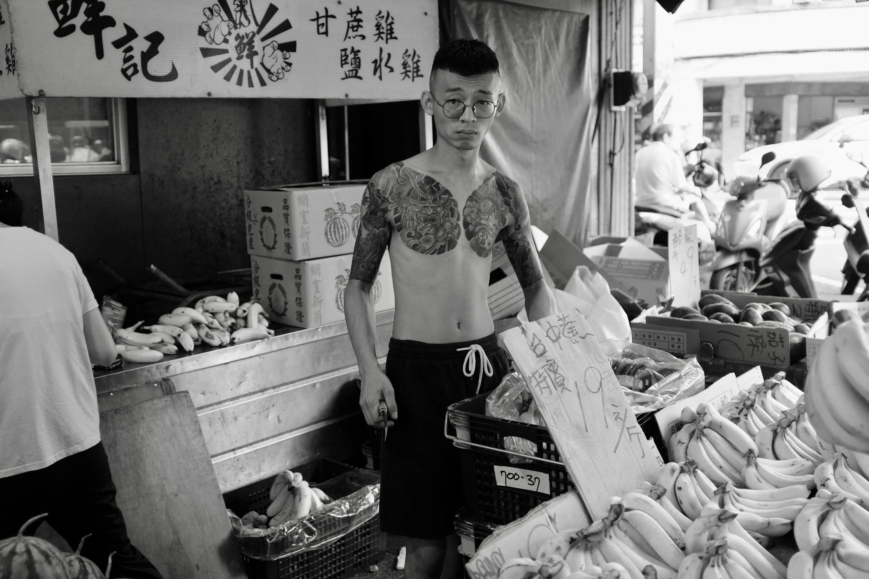 Black and white street photograph of a tattooed man standing among bananas and handwritten signs at a fruit stall