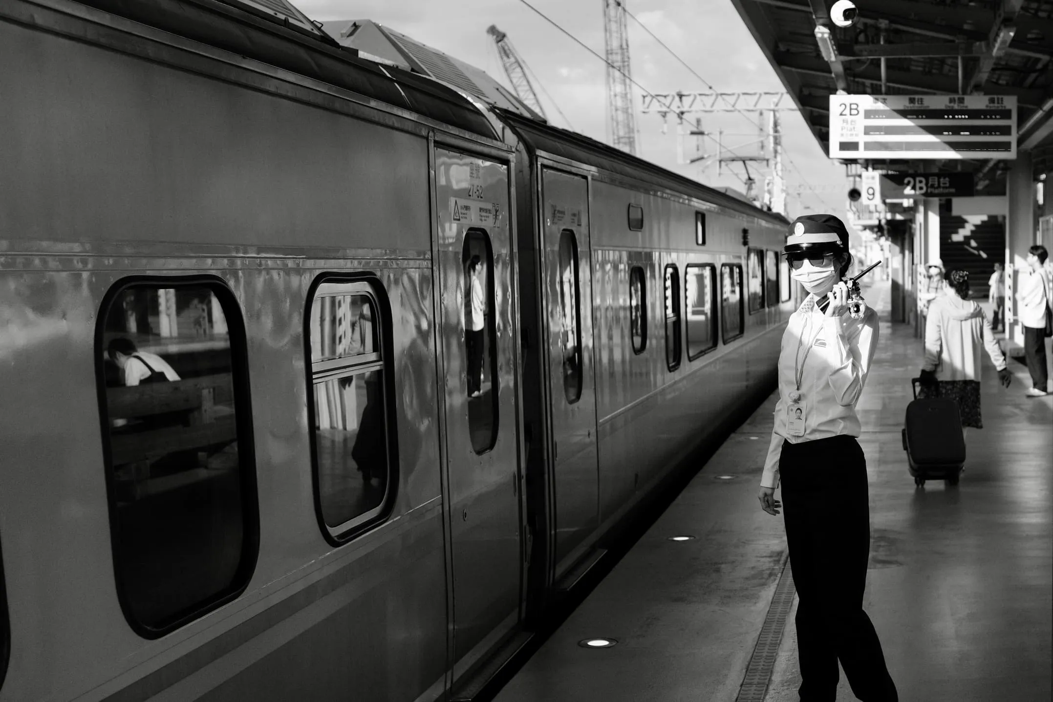 Black and white street photograph of a train platform attendant with passengers and luggage beside a train