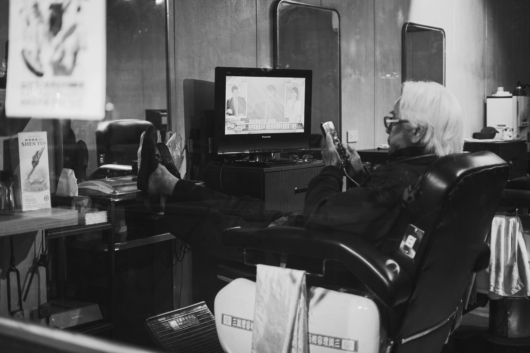Black and white view through an East District barbershop window of a white-haired person reclining in a chair while looking at a phone beside a television in Taipei