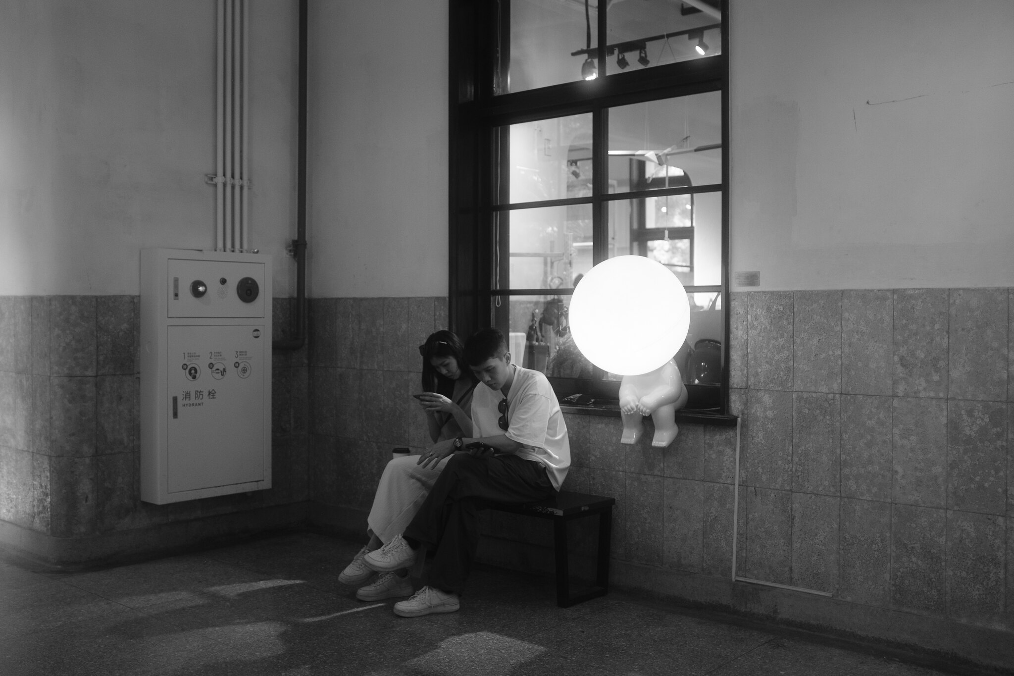 Black and white street photograph of a couple checking phones beneath a glowing window display in Taipei's East District