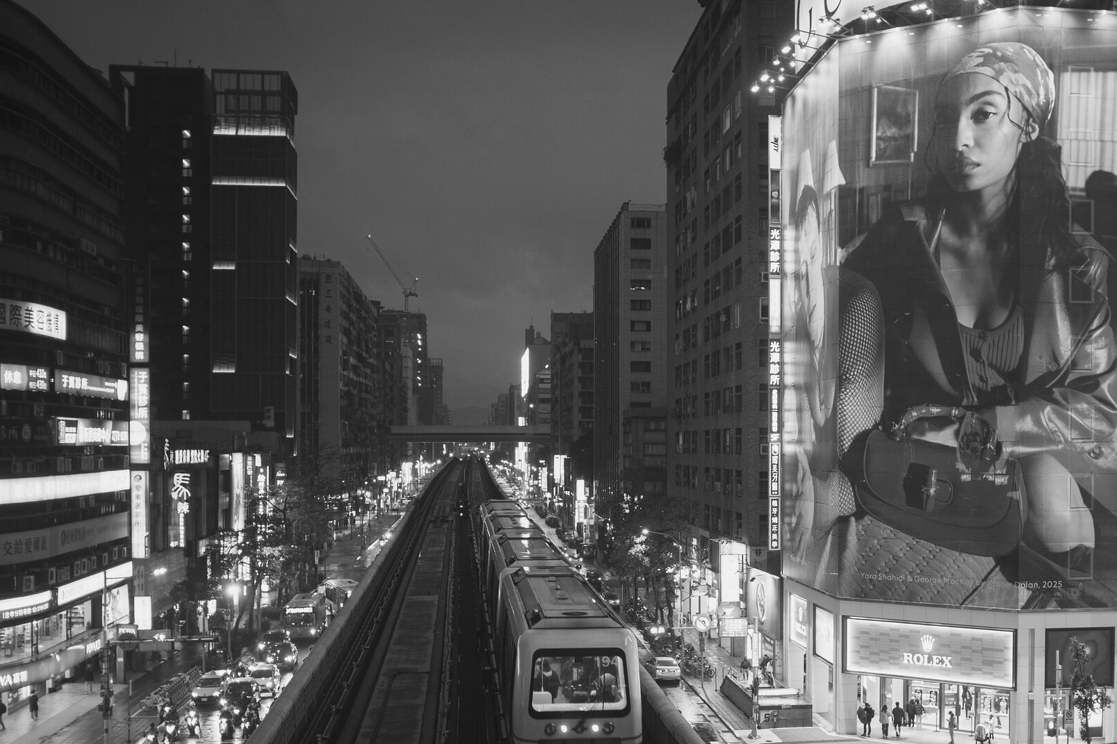 Black and white view of a Taipei Metro train entering the East District at night beside a towering fashion billboard and illuminated storefronts