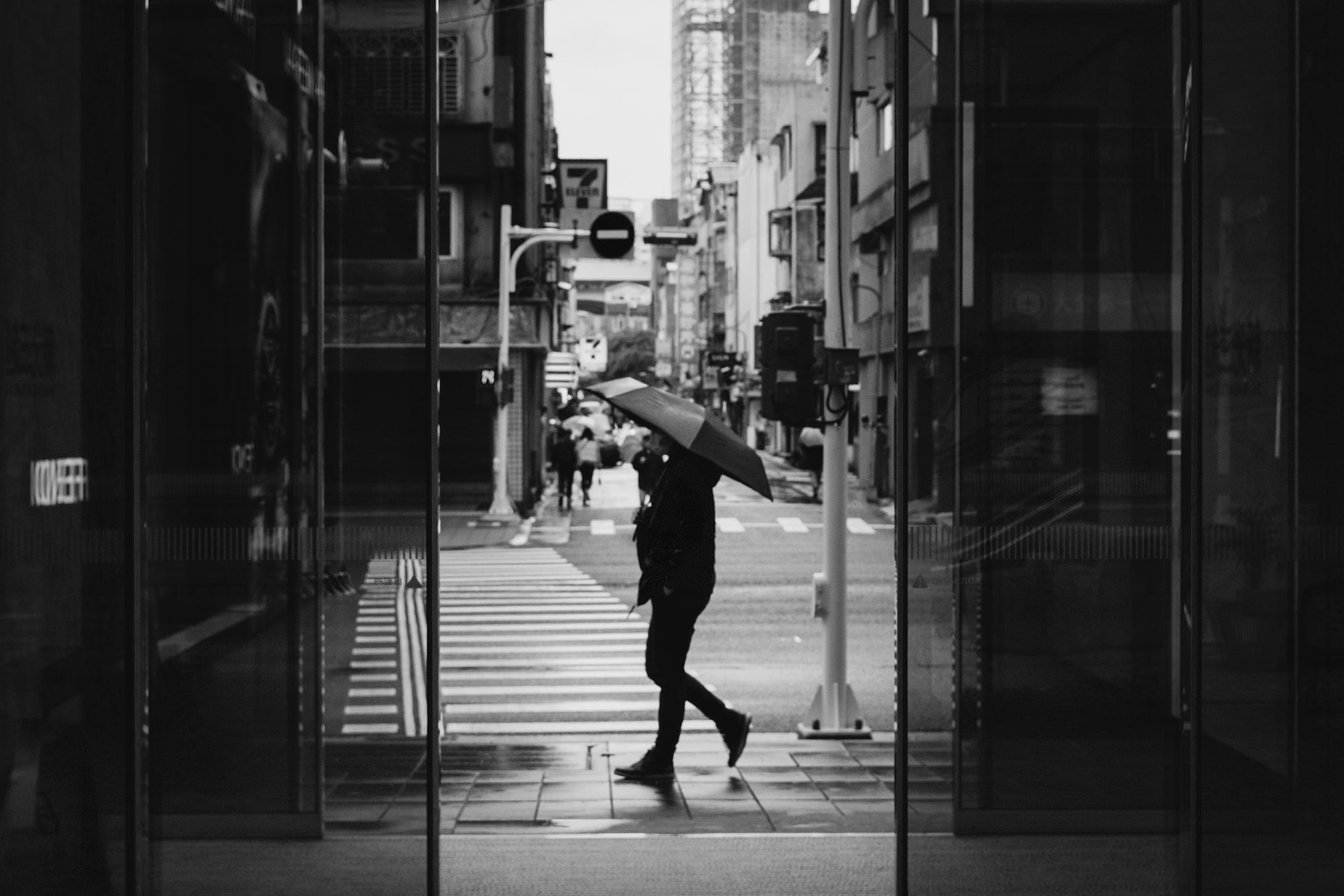Black and white street photo of a person carrying an umbrella across a rainy East District crosswalk, framed by glass reflections in Taipei