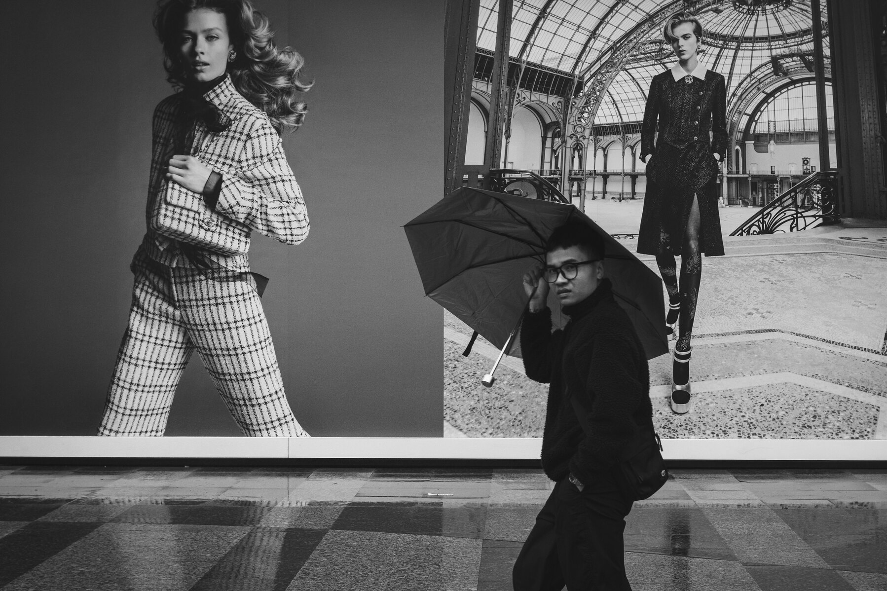Black and white street photograph of a person carrying an umbrella past oversized fashion advertisements on a rainy East District sidewalk in Taipei
