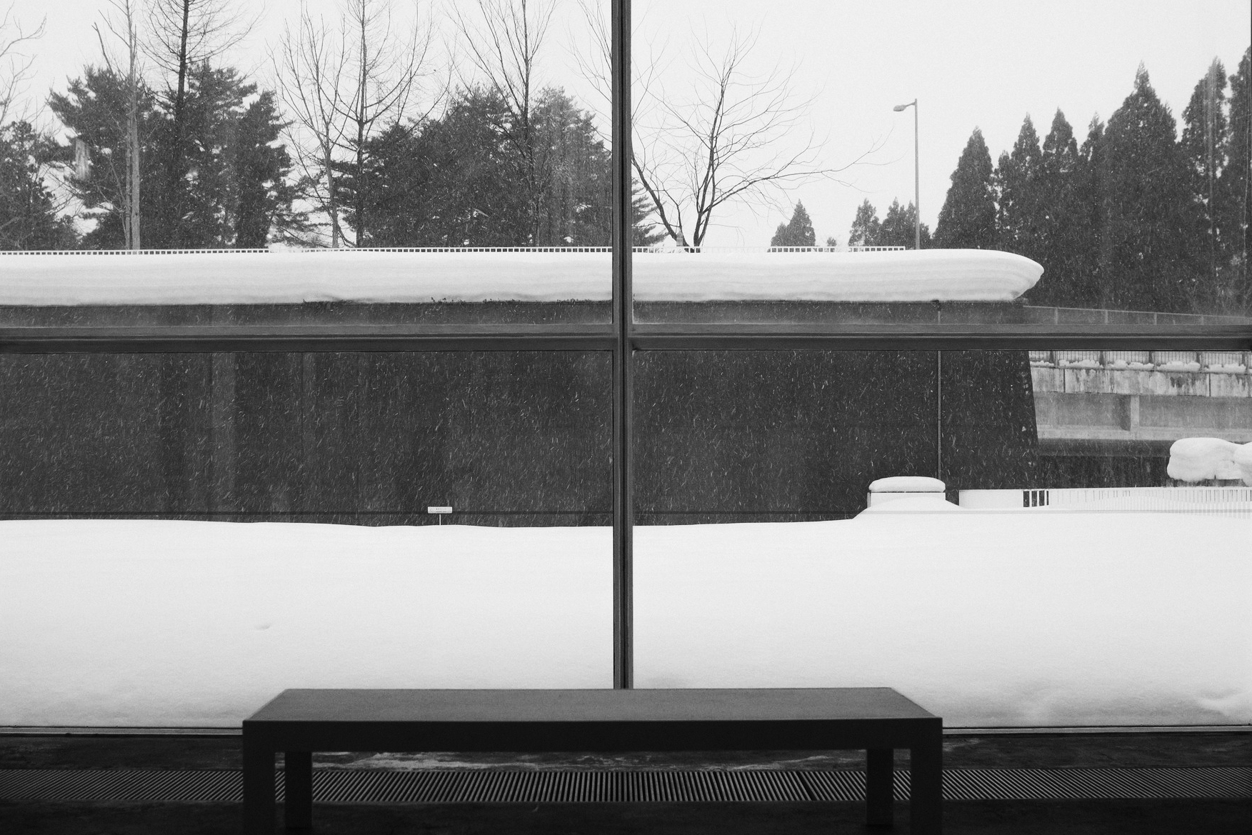 Black and white snowy landscape seen through a window, with a dark bench in the foreground