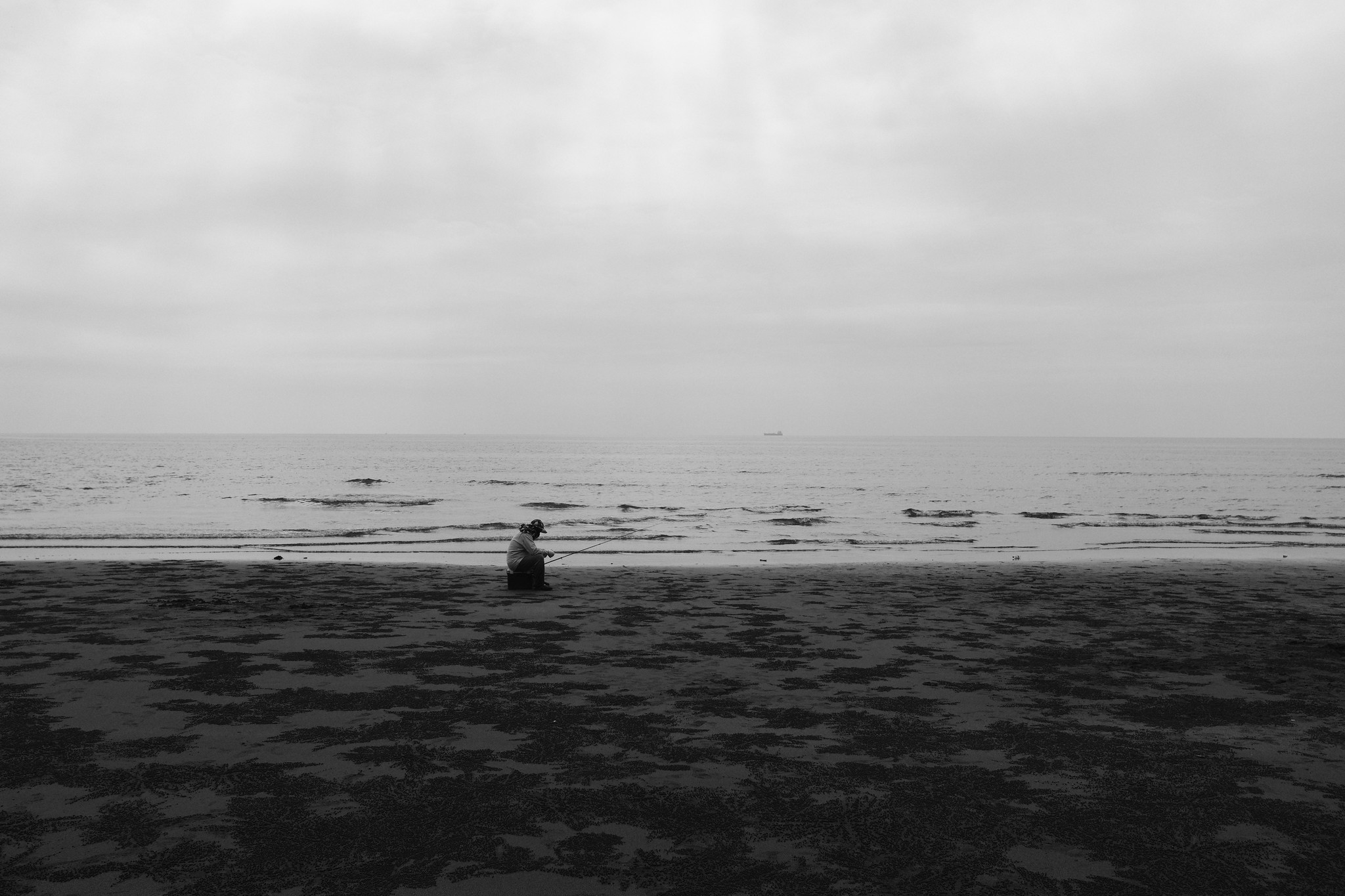 Black and white intertidal coast scene with a lone fisherman seated on the shore under a wide pale sky
