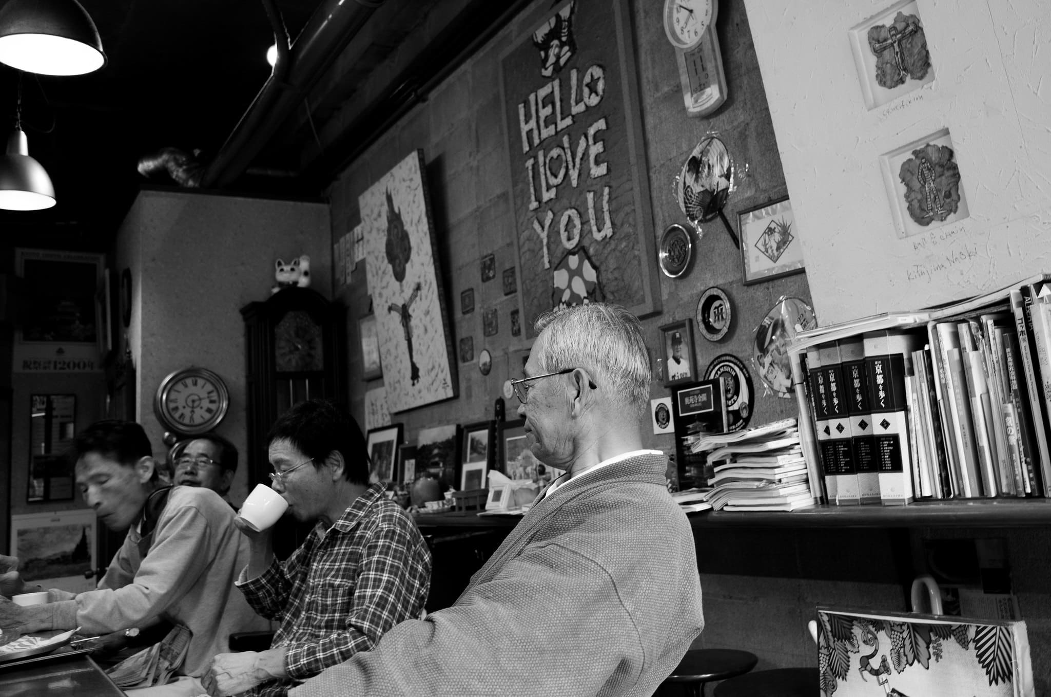 Black and white cafe interior in Japan with older patrons seated beneath wall art, books and clocks