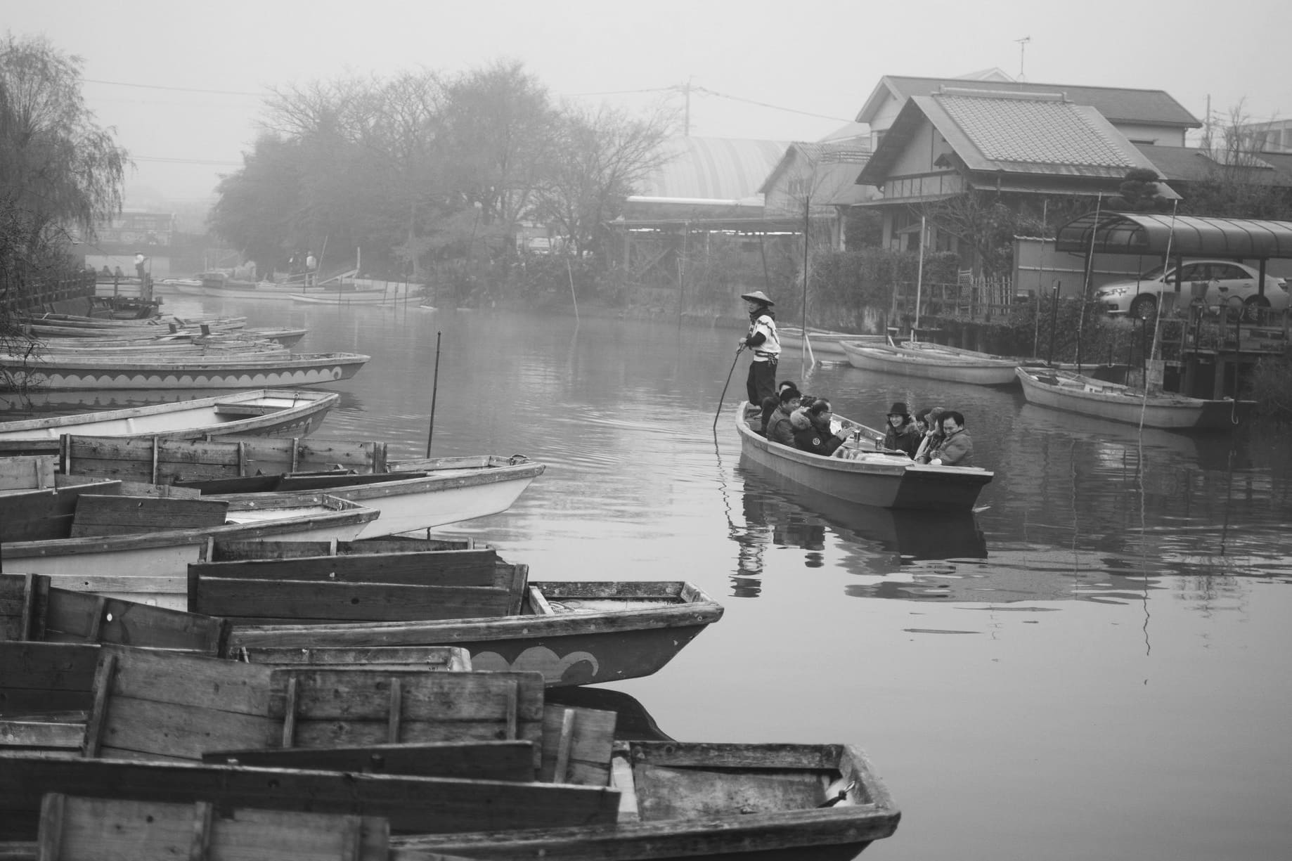 Black and white canal scene in Japan with a boat operator guiding passengers between moored wooden boats