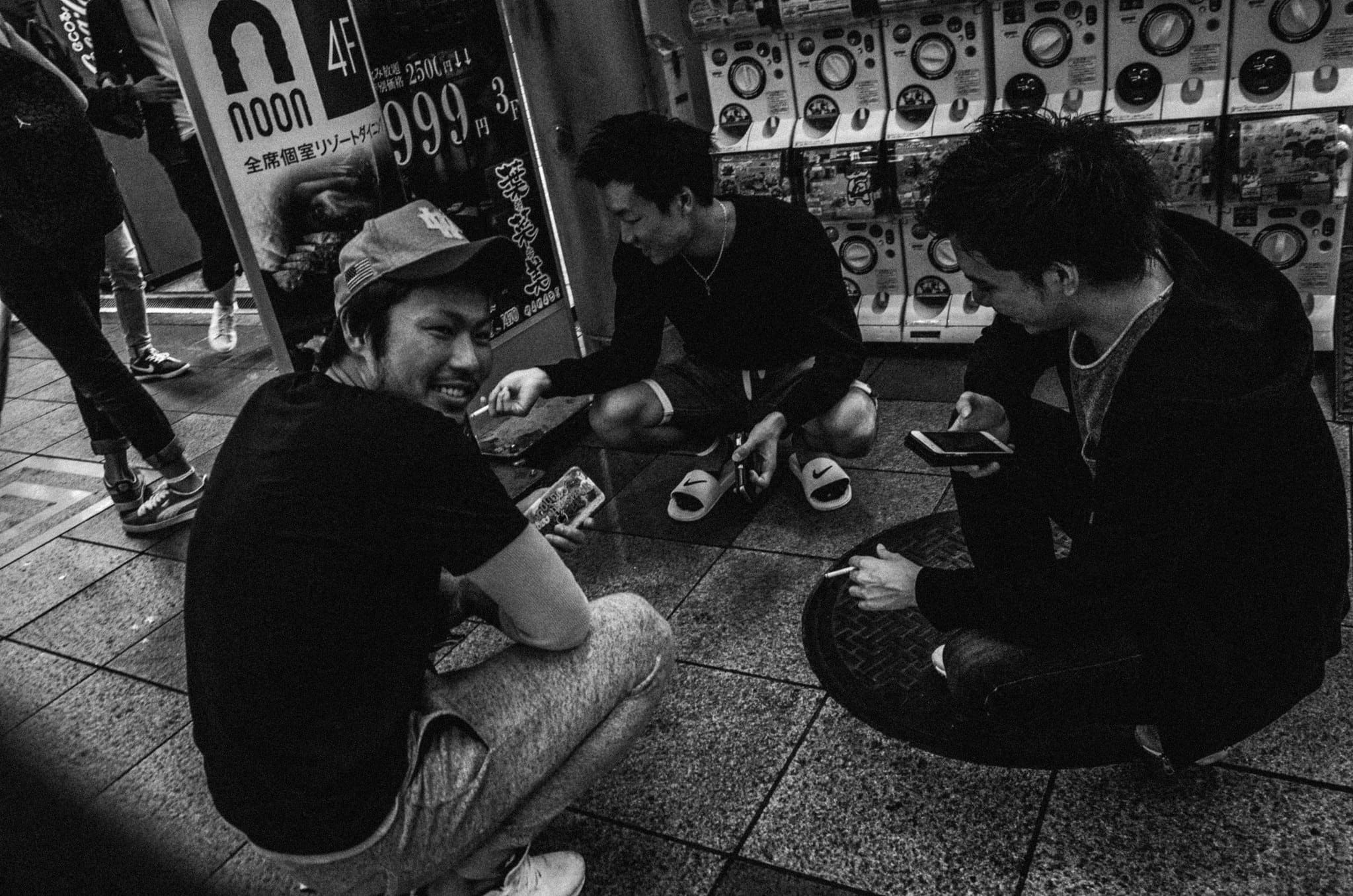 Black and white night street in Japan with friends crouching beside rows of capsule toy machines