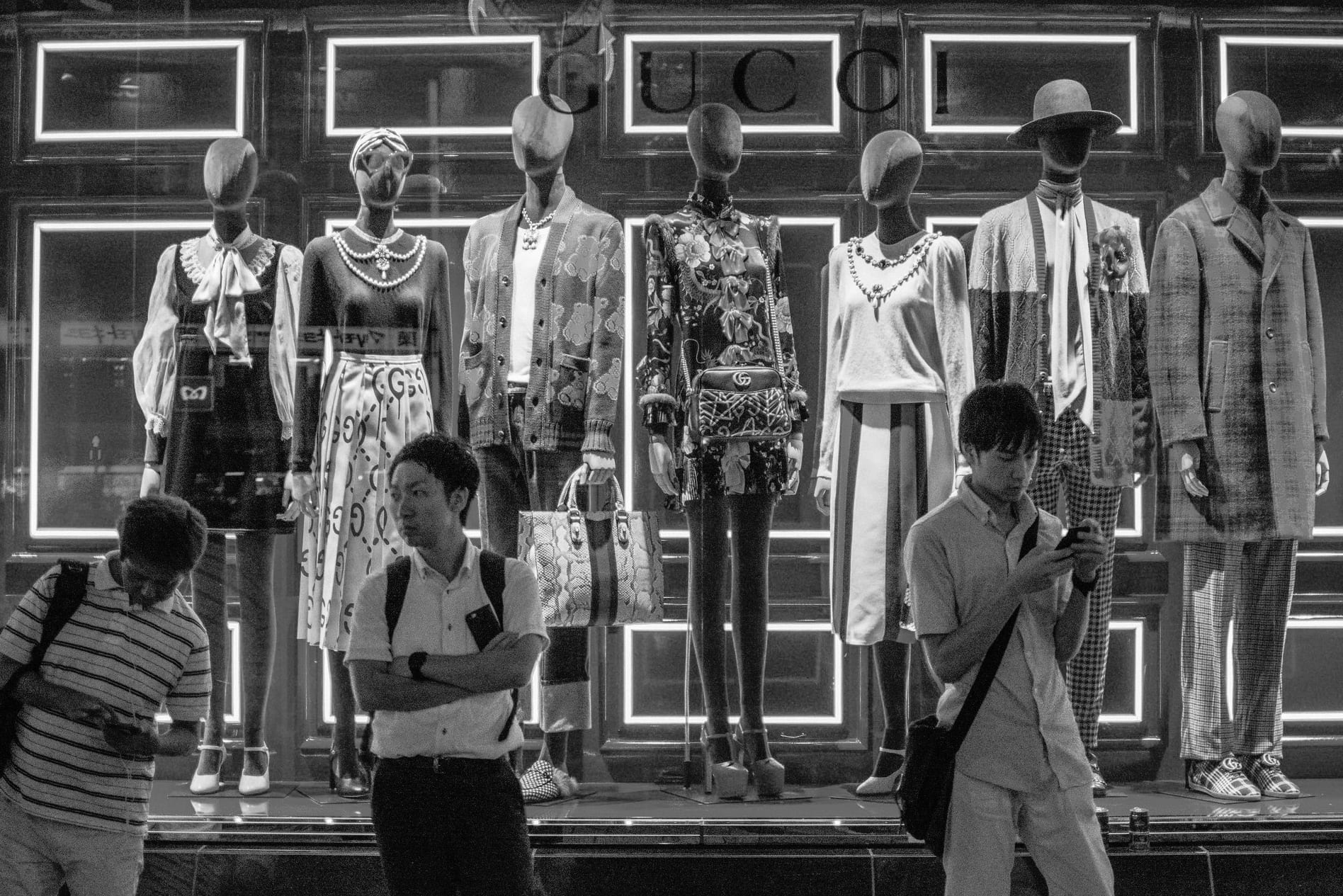 Black and white Japan storefront with commuters standing before an illuminated fashion display of mannequins