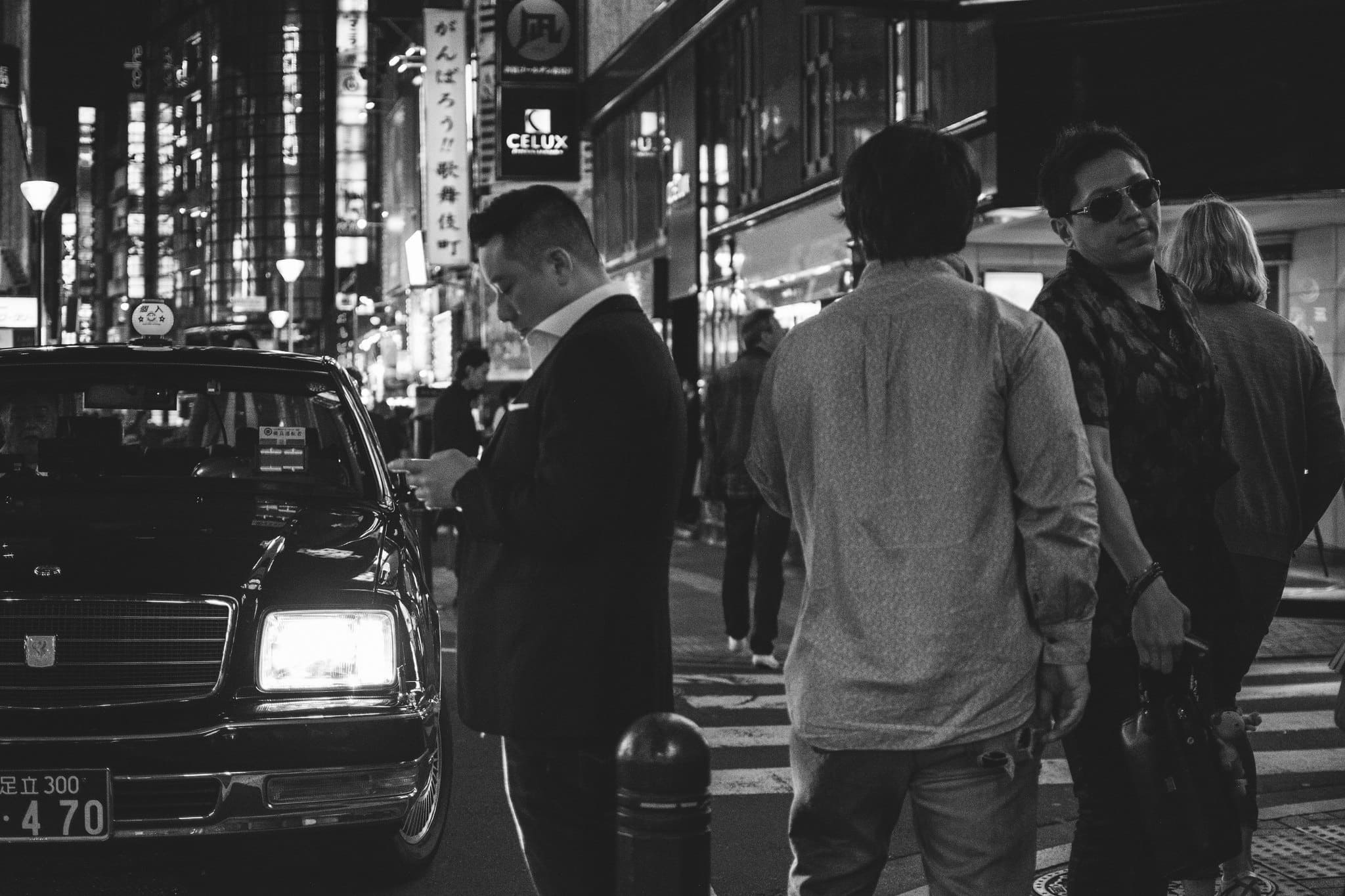 Black and white night street in Japan with a parked taxi and pedestrians gathered beneath illuminated signs