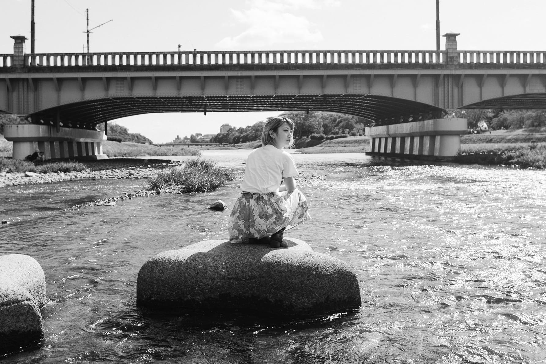 Black and white portrait of a woman seated on a stepping stone in a Japanese river beneath a bridge