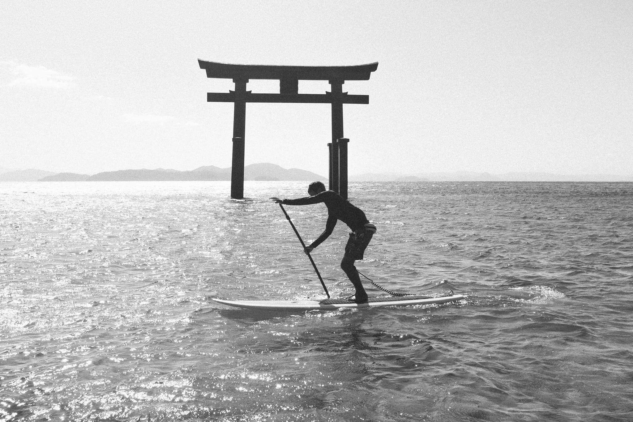 Black and white silhouette of a paddleboarder crossing sunlit water before a torii gate in Japan