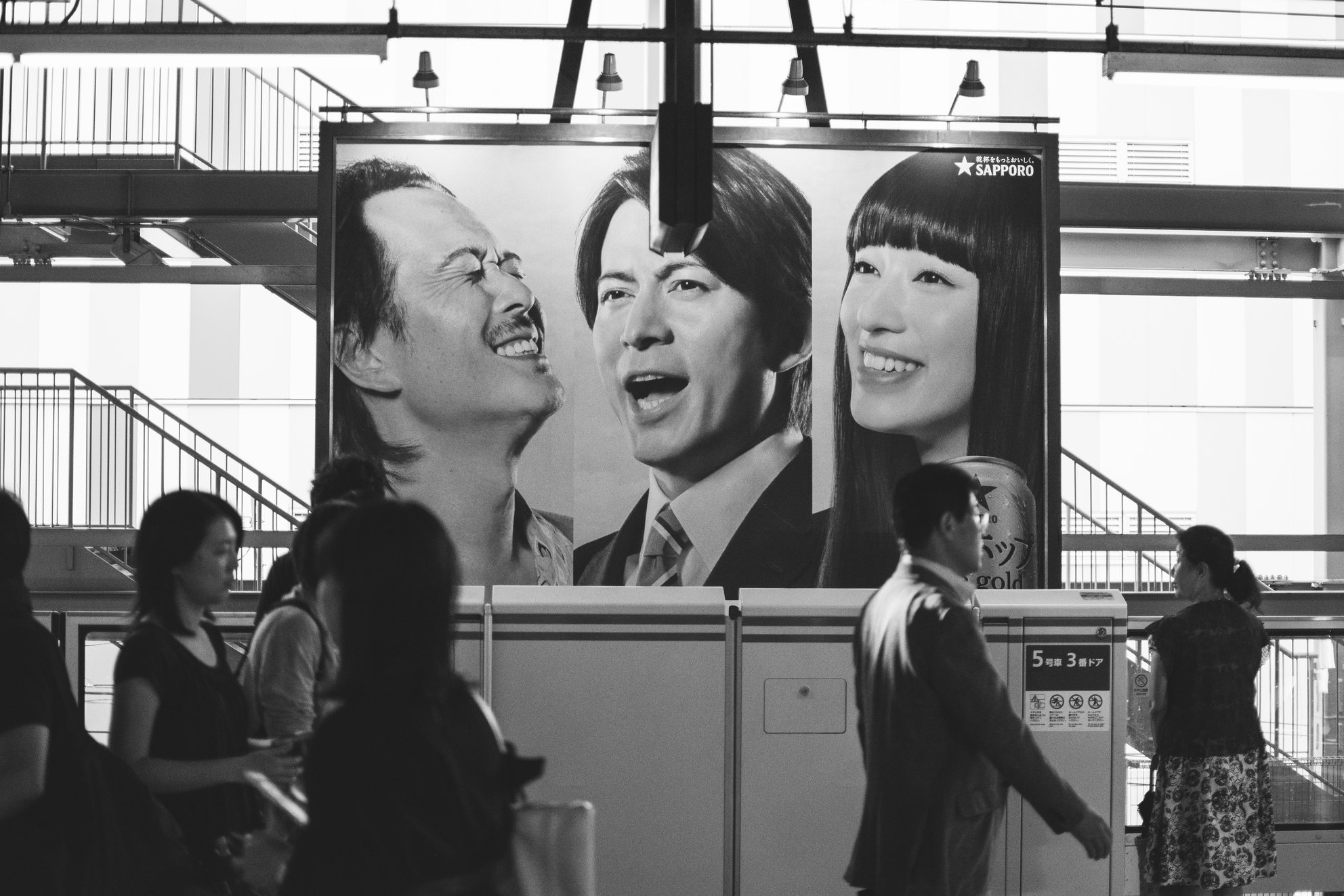 Black and white station scene in Japan with passersby moving beneath large portrait advertisements