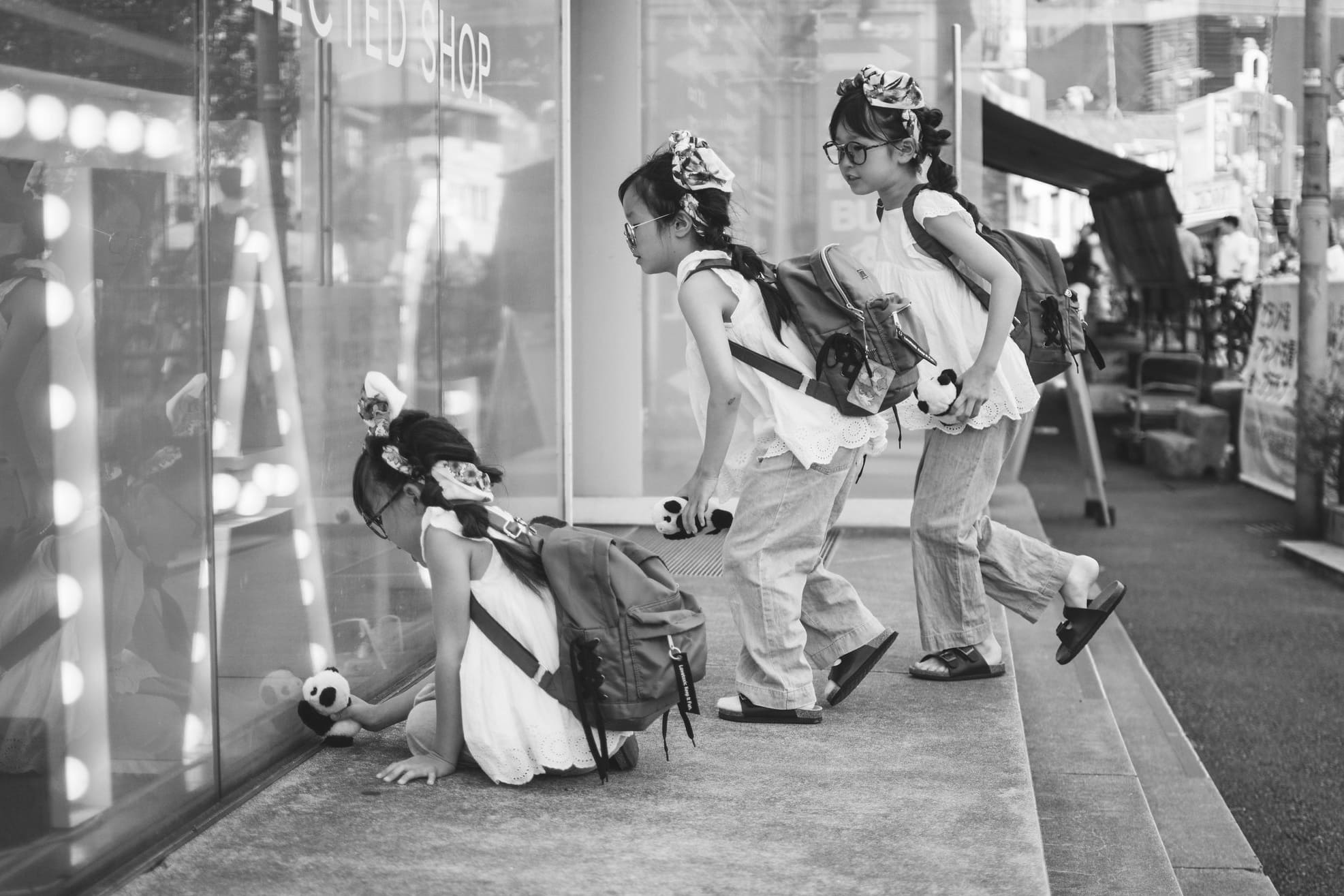 Black and white street scene in Japan with three girls peering into a shop window while holding panda toys