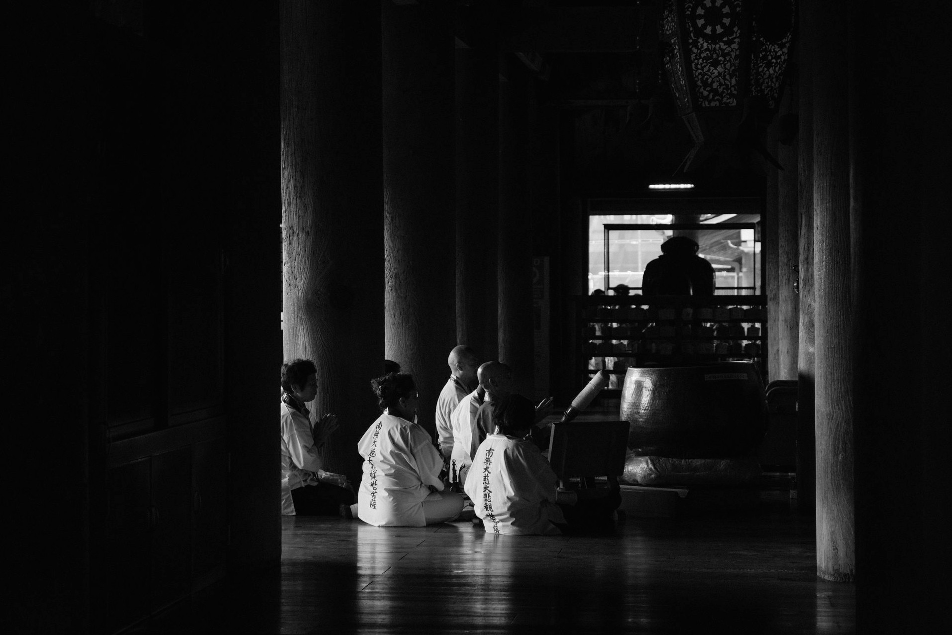 Black and white scene in a Japanese temple with pilgrims in white robes kneeling in prayer between wooden pillars