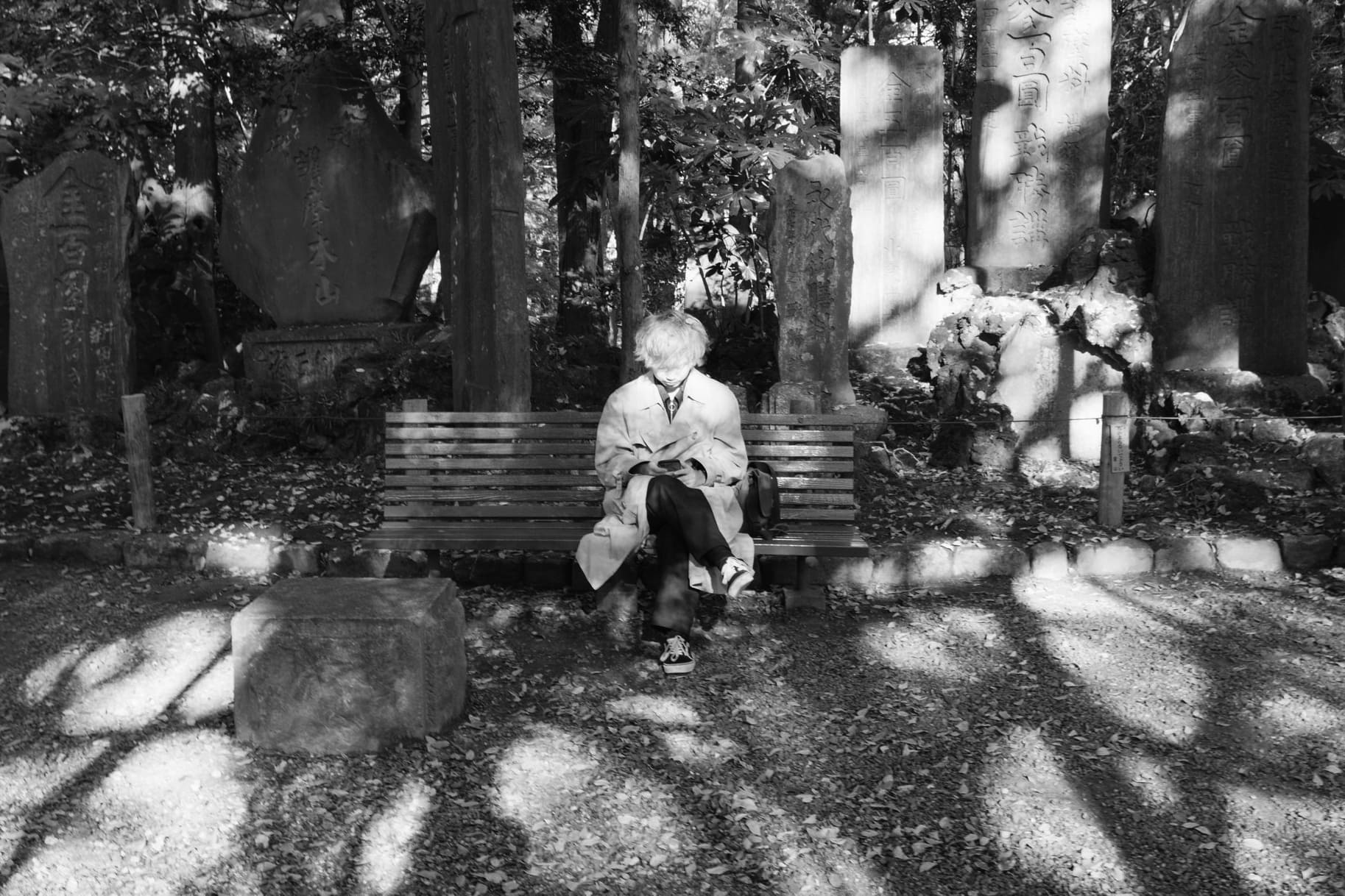 Black and white scene in Japan with a person checking a phone on a bench among stone monuments