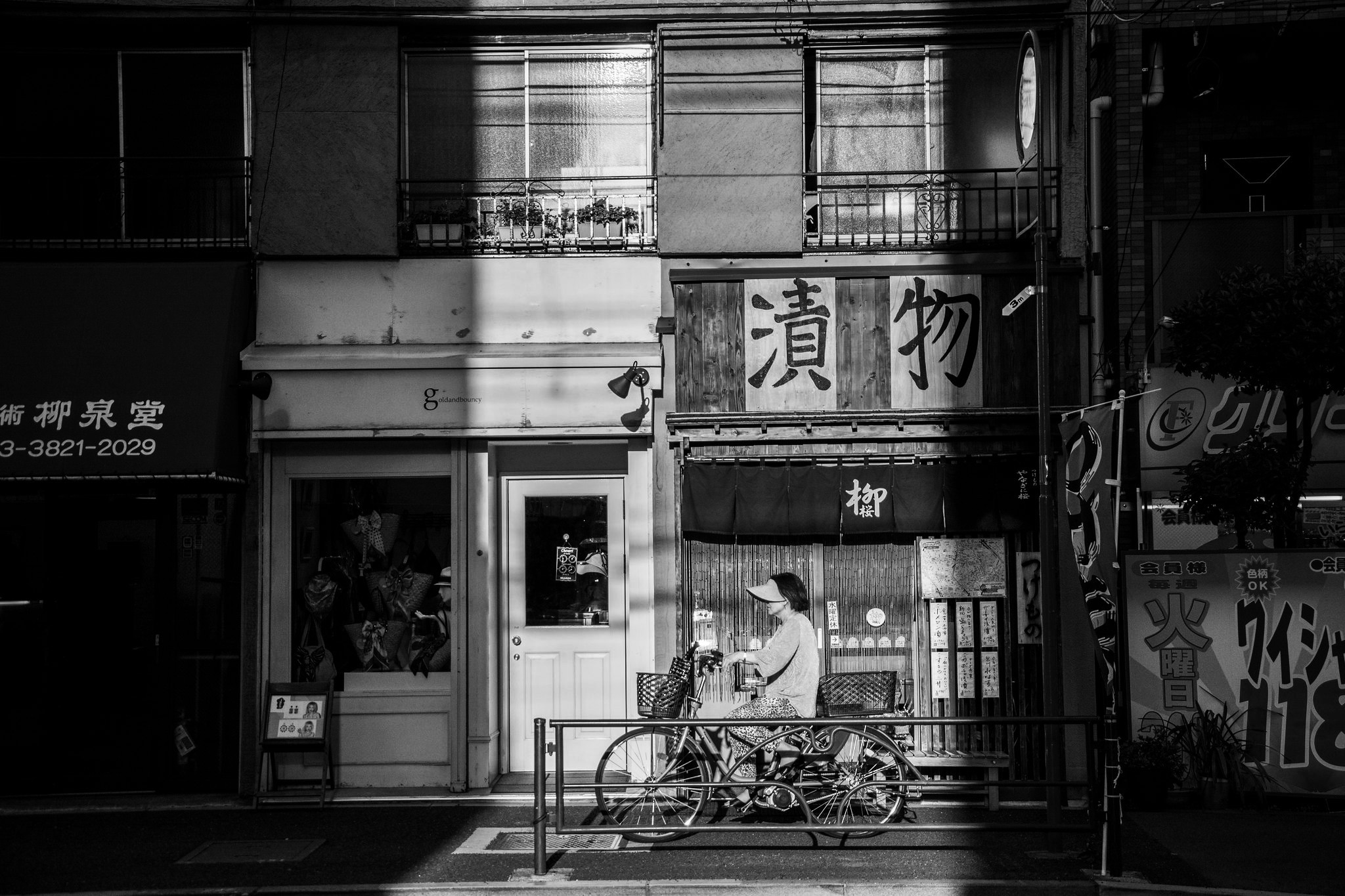 Black and white Japanese street scene with a cyclist passing a wooden shopfront with traditional tsukemono signage