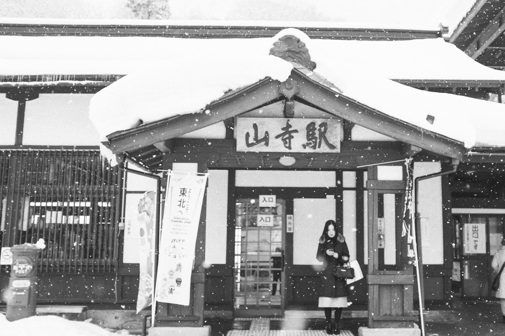 Black and white winter scene with a woman standing outside snow-covered Yamadera Station in Japan