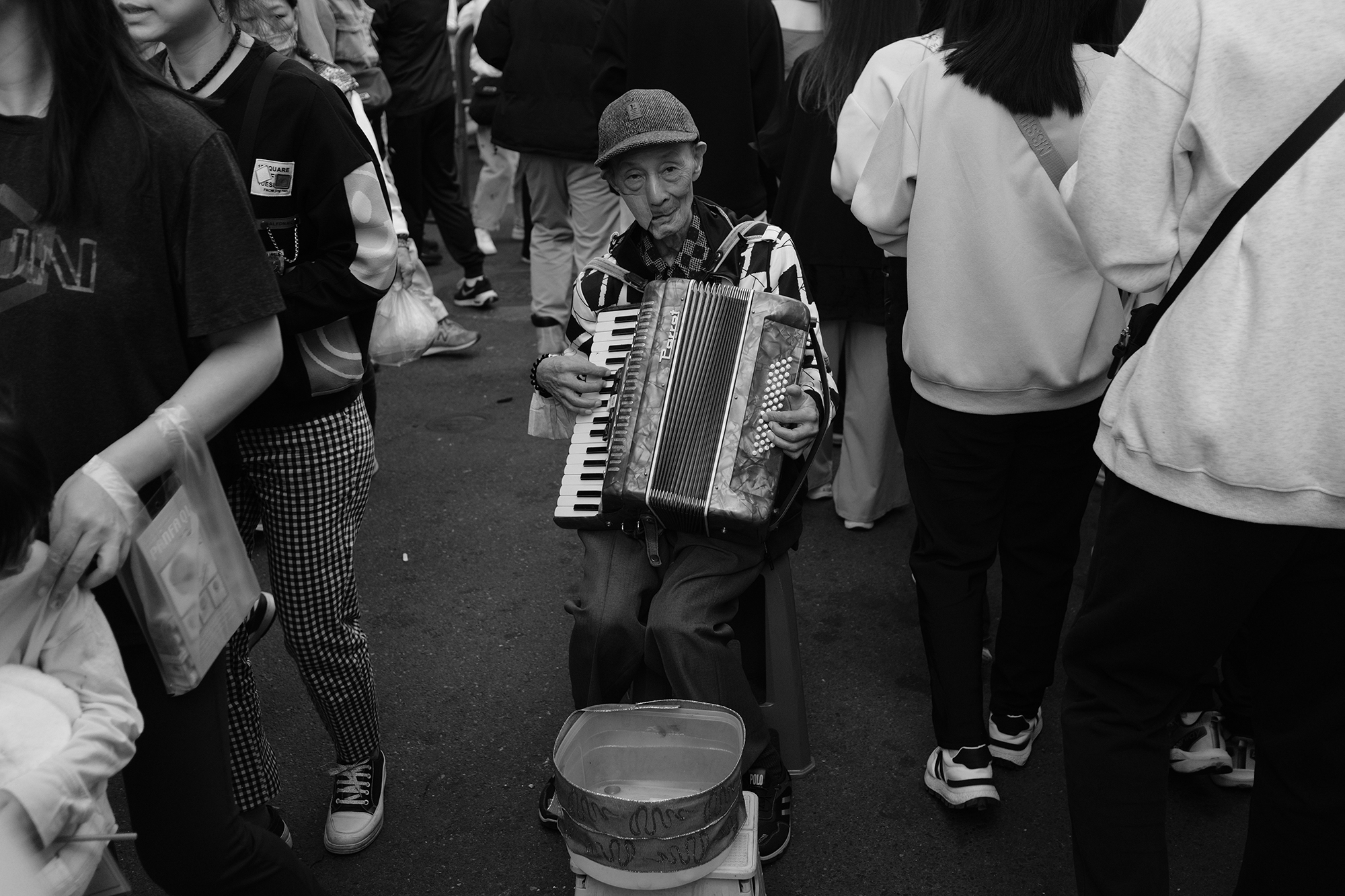Candid black and white street photo of an elderly accordion player surrounded by a crowd in Keelung, Taiwan