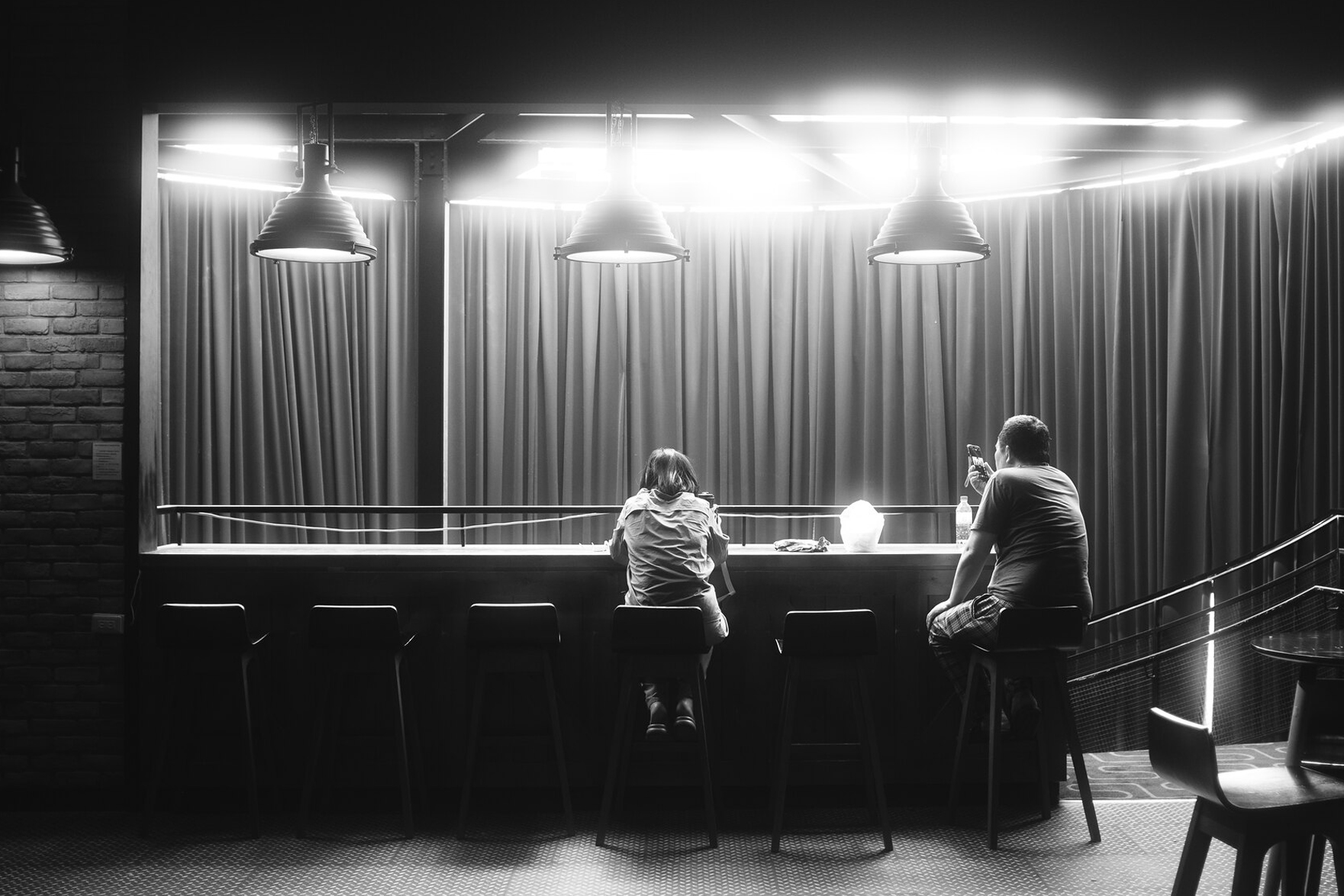 Black and white street interior scene of two people seated at a bar counter under bright lamps, framed by curtains and layered night light in Taiwan