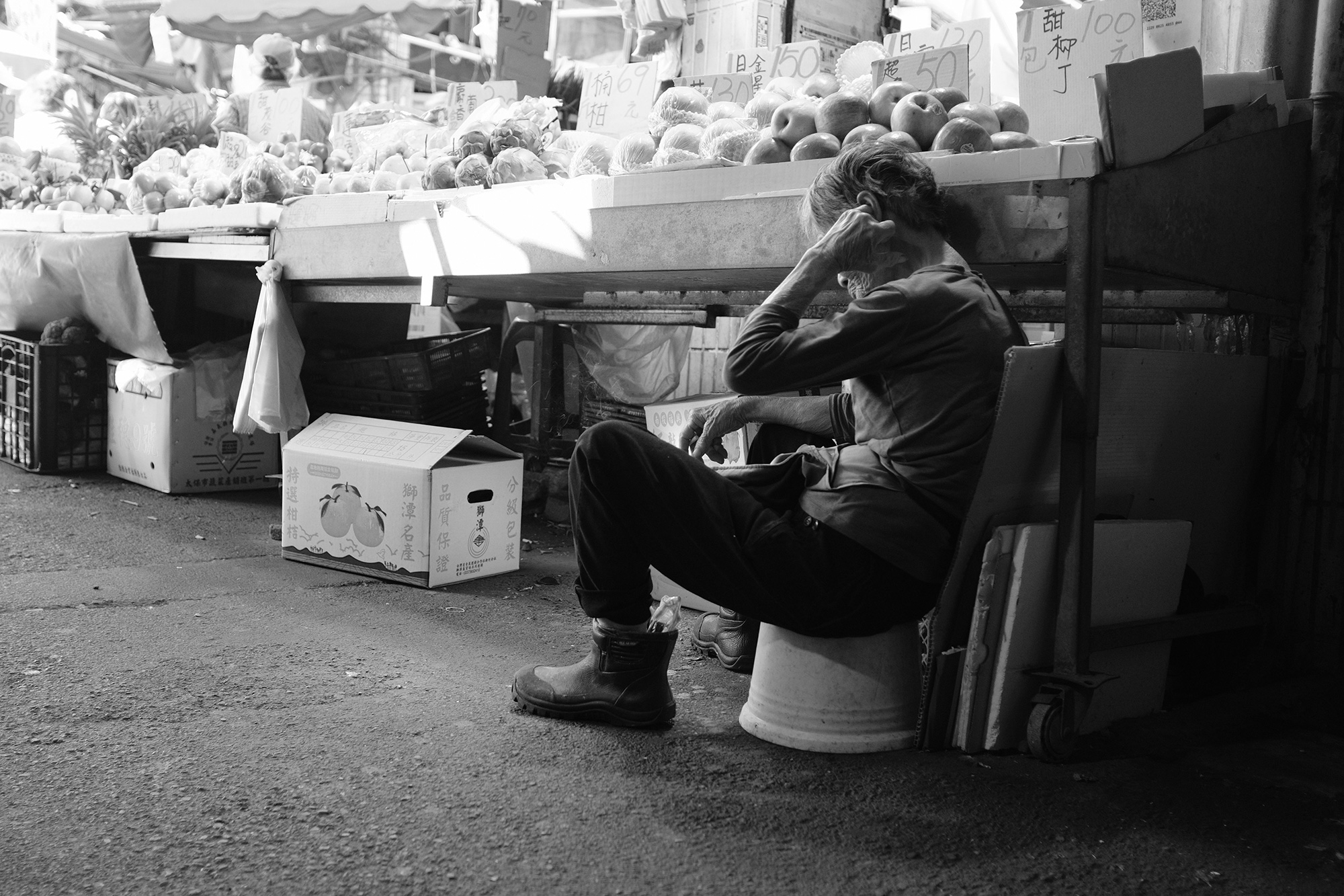 Black and white street photograph of a market vendor resting beneath a fruit stand, framed by layered urban textures in Taiwan