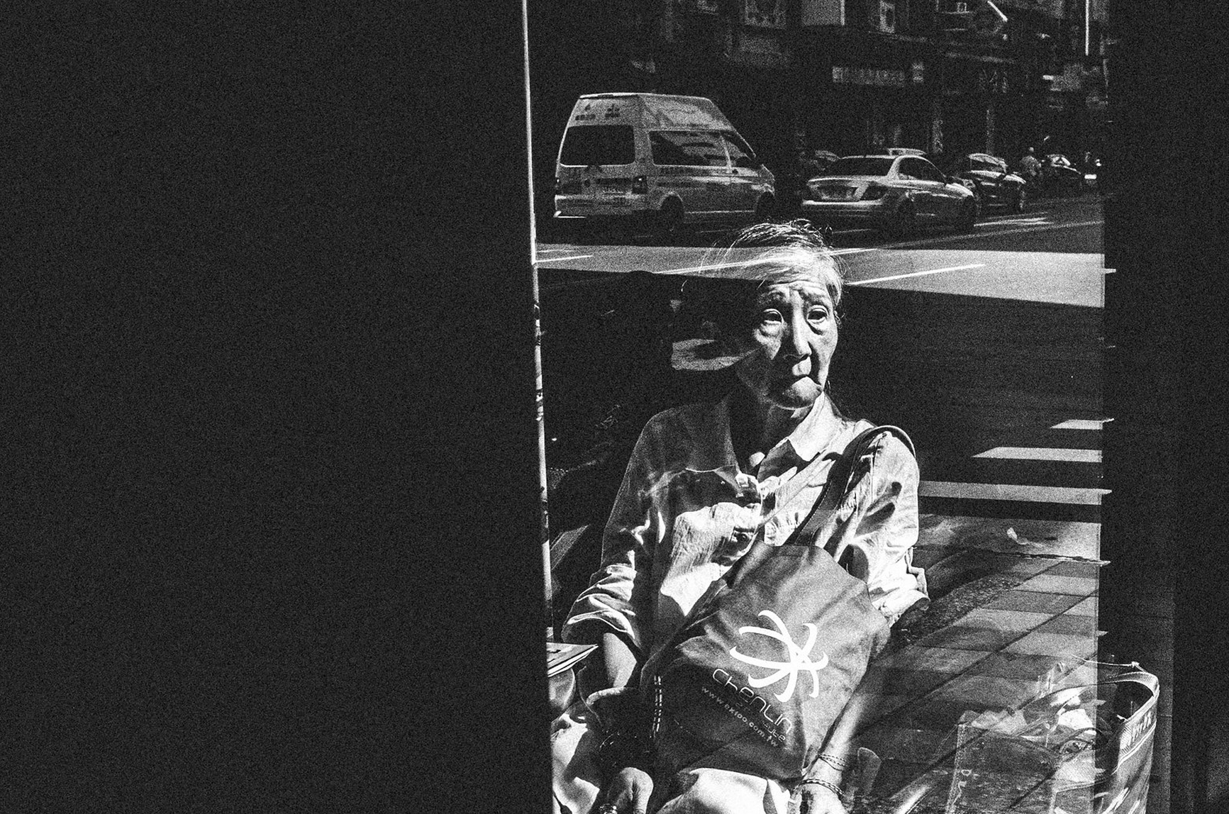 High-contrast black and white street photo of an elderly woman seen through a window reflection