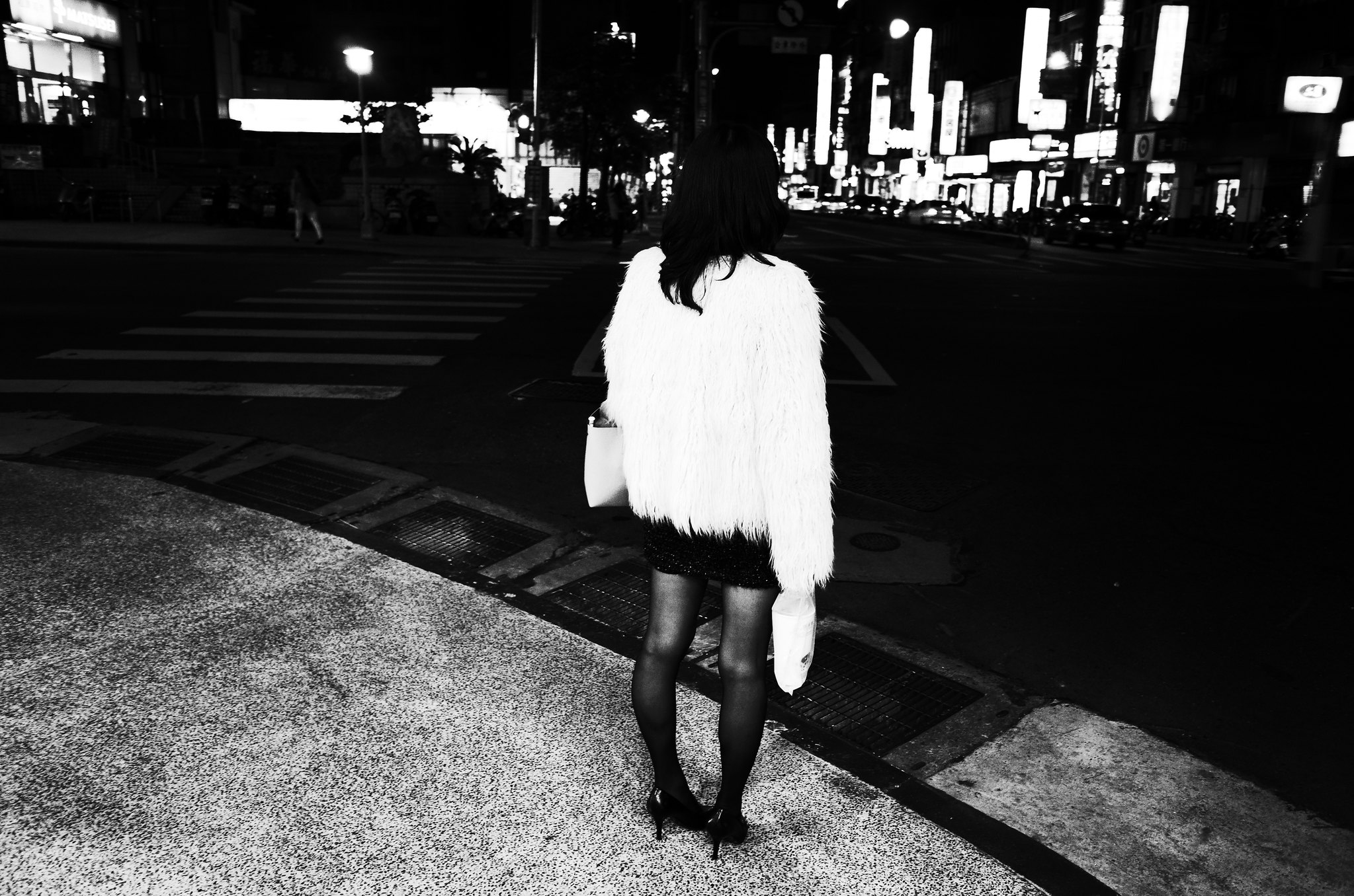 Black and white street photograph of a woman from behind, wearing a furry coat at a city intersection at night