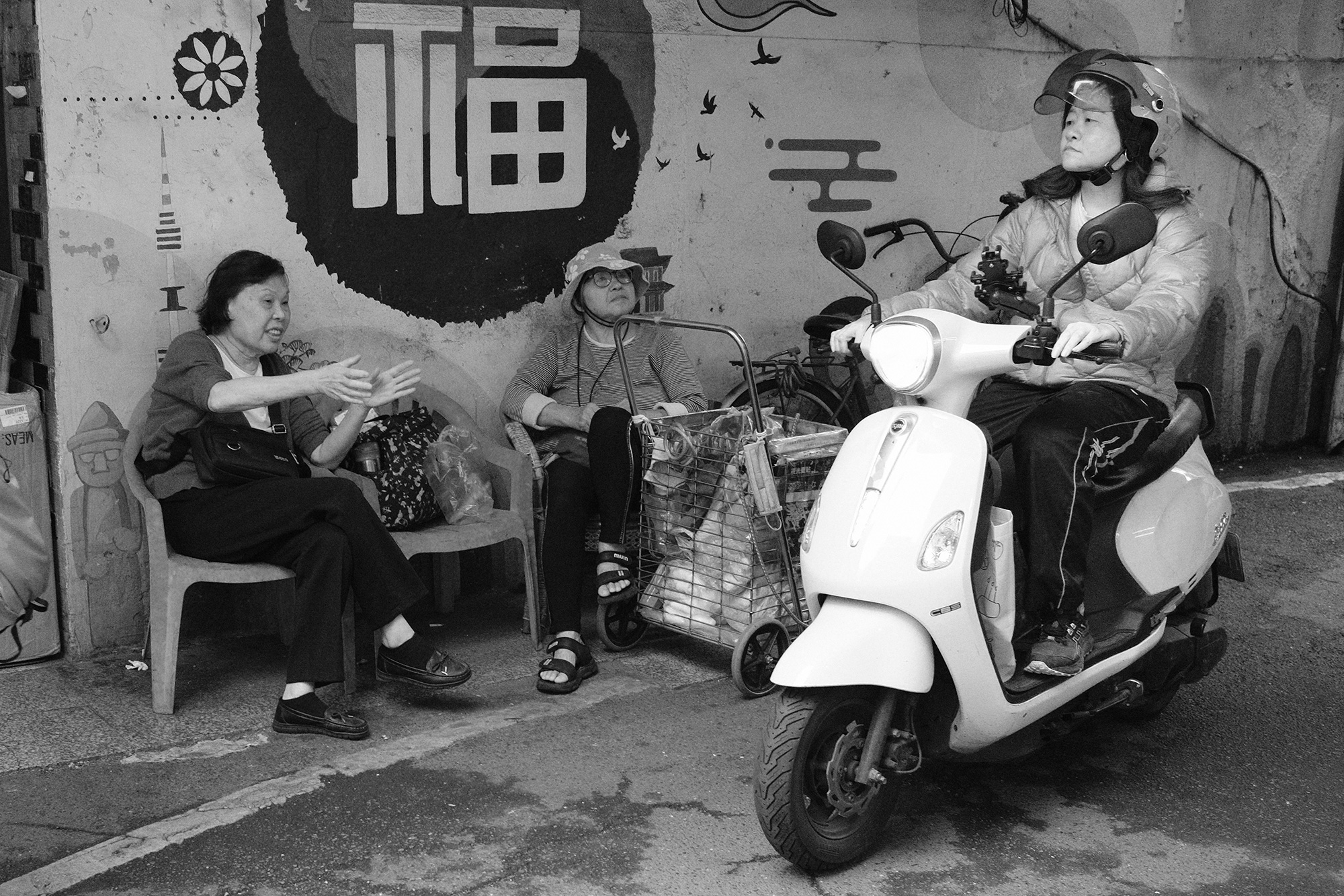 Black and white street scene of neighborhood conversation beside a scooter, showing everyday urban layers in Taiwan