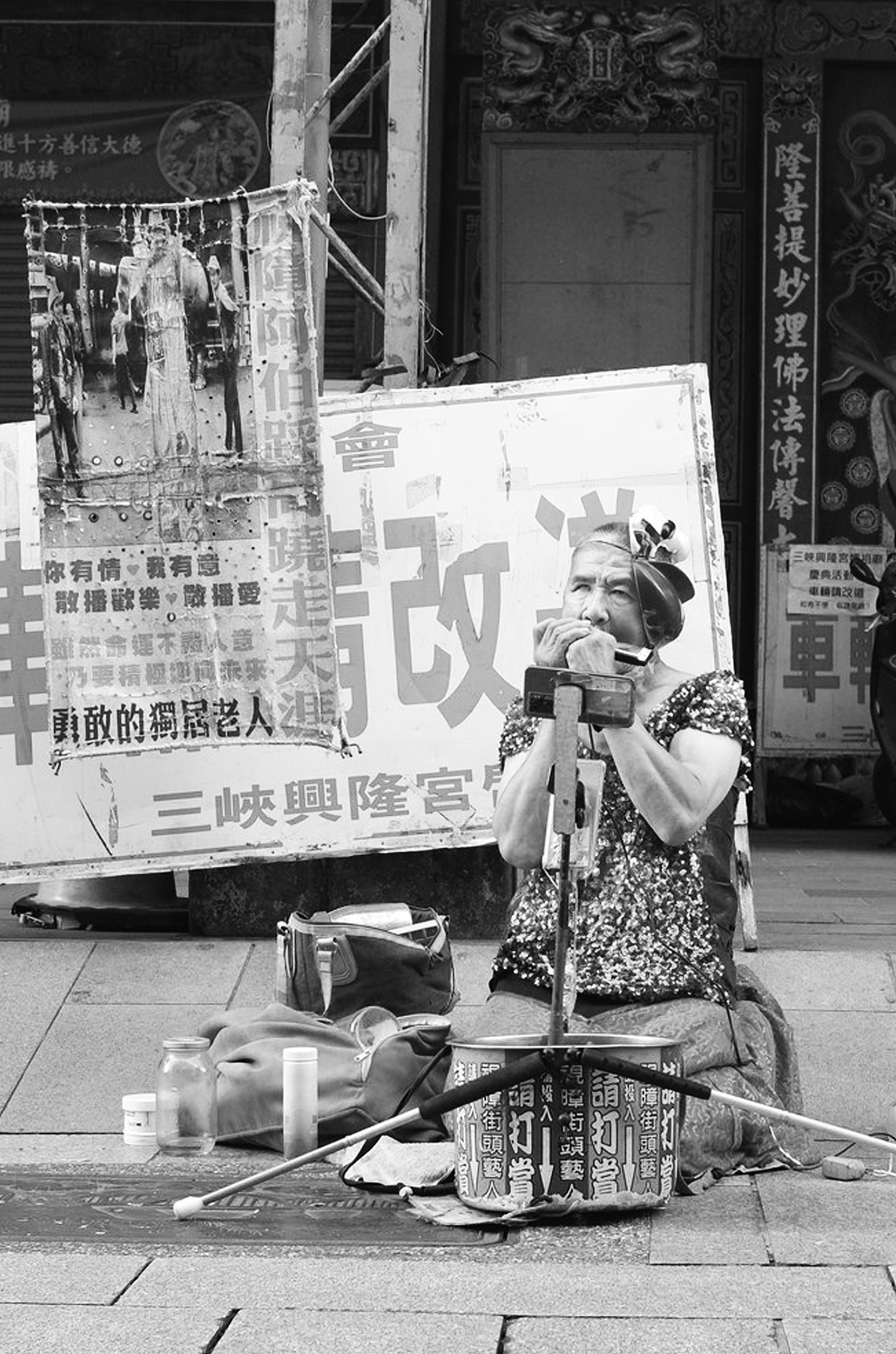 Black and white vertical street photograph of a seated man beside a large hand-lettered sign outside a temple entrance