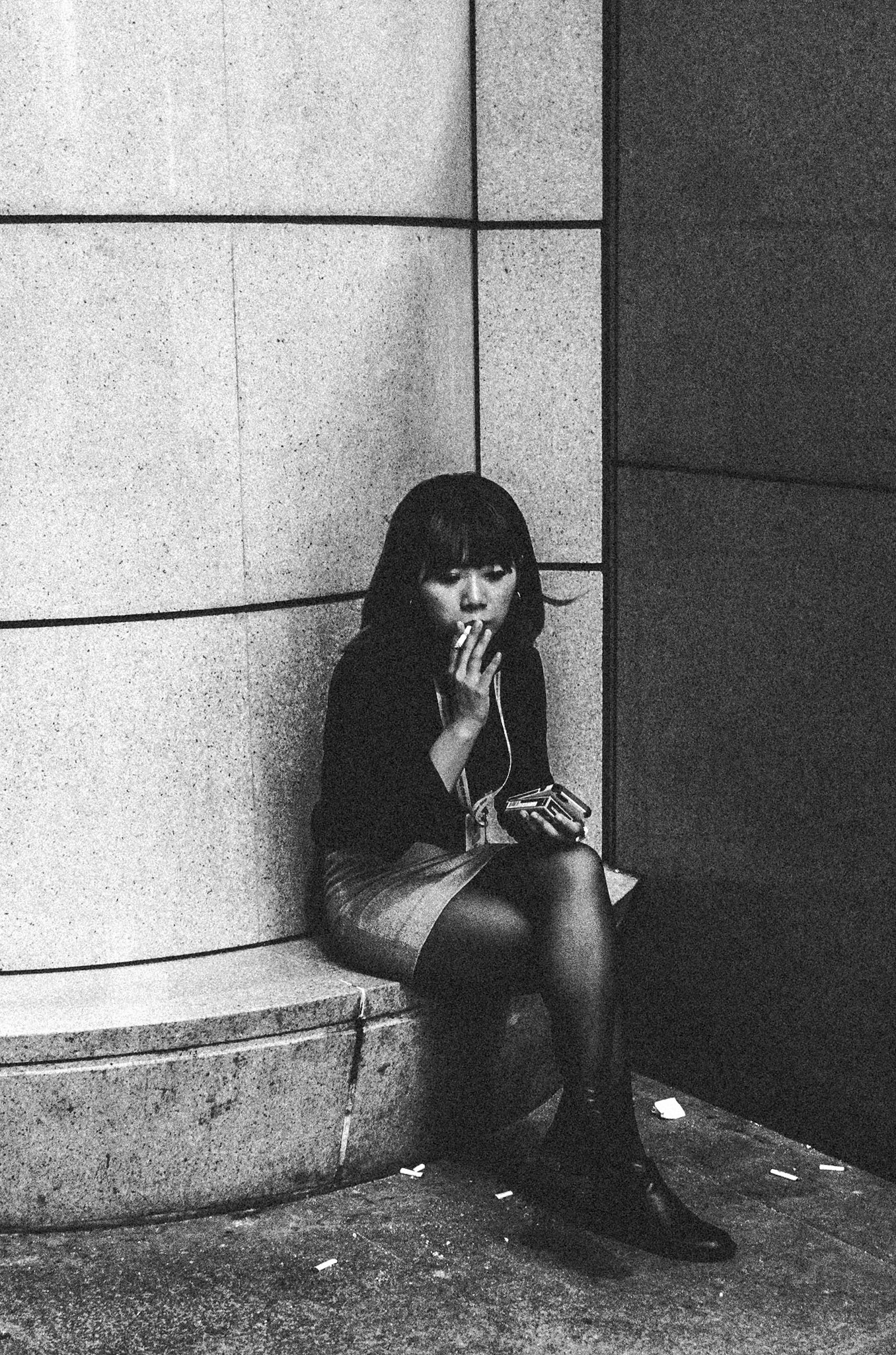 Black and white portrait of a young woman with earphones leaning against a concrete wall while smoking