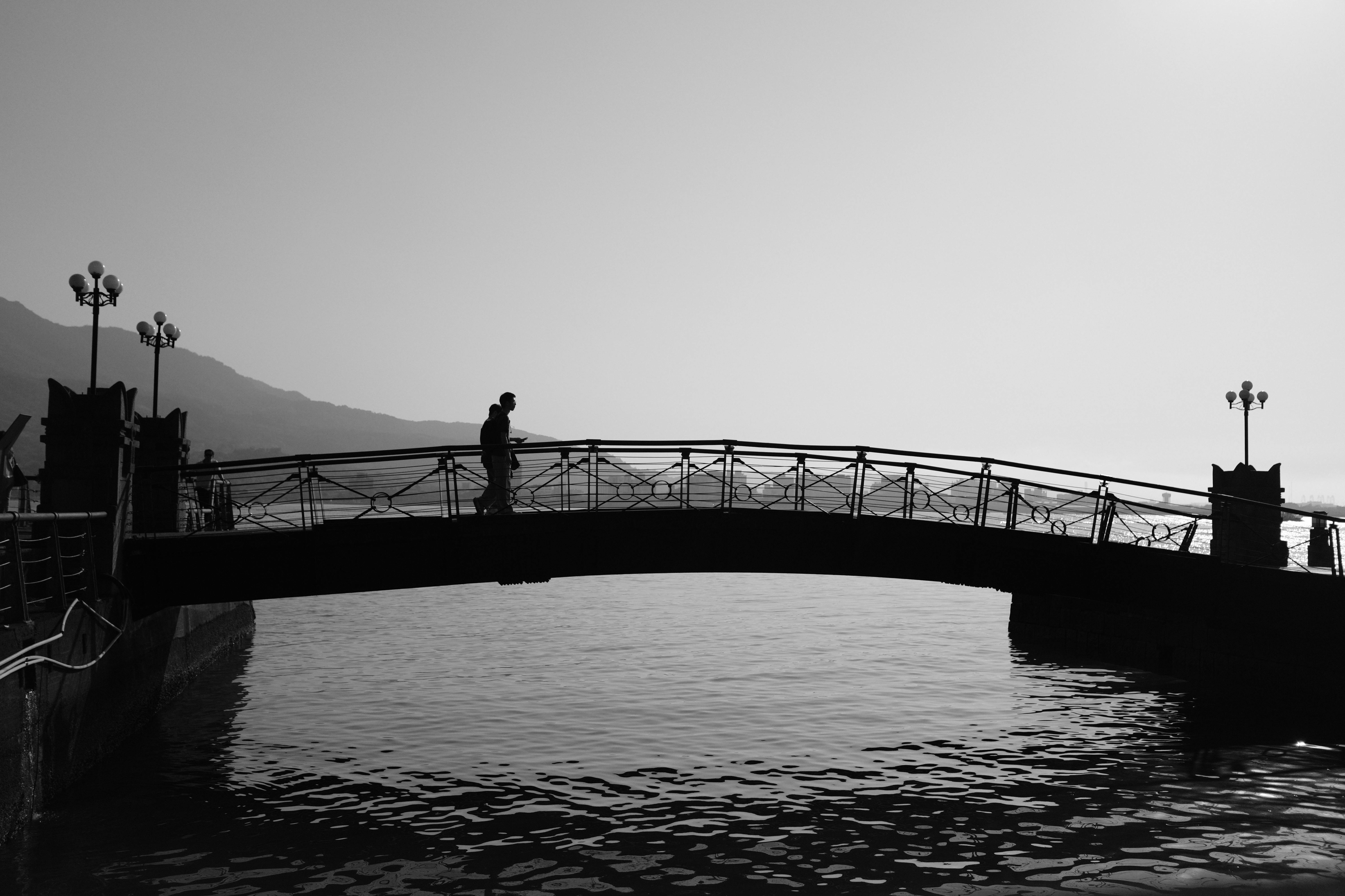 Minimal black and white street photo of pedestrians crossing a silhouetted bridge along the Tamsui waterfront in Taipei