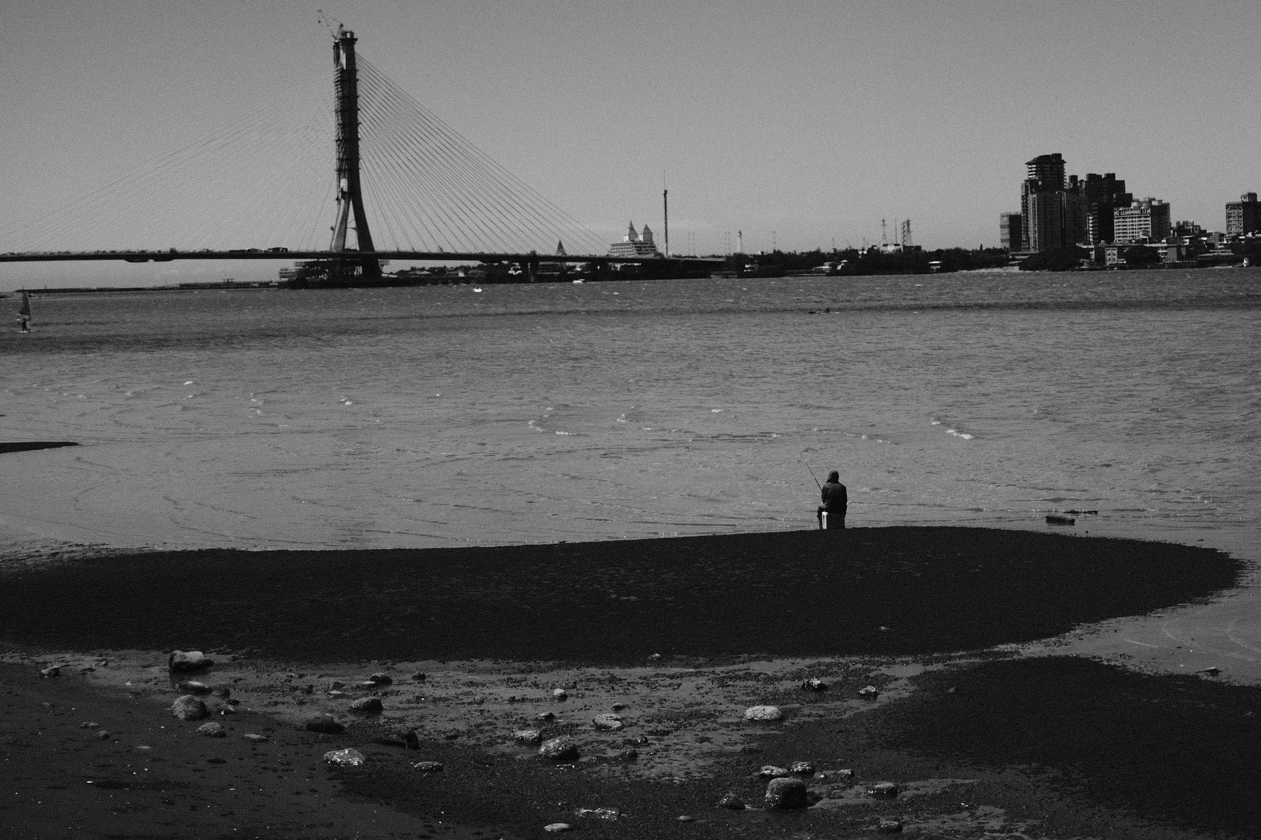 Quiet black and white riverside scene with a lone figure facing the bridge and skyline in Tamsui, Taipei