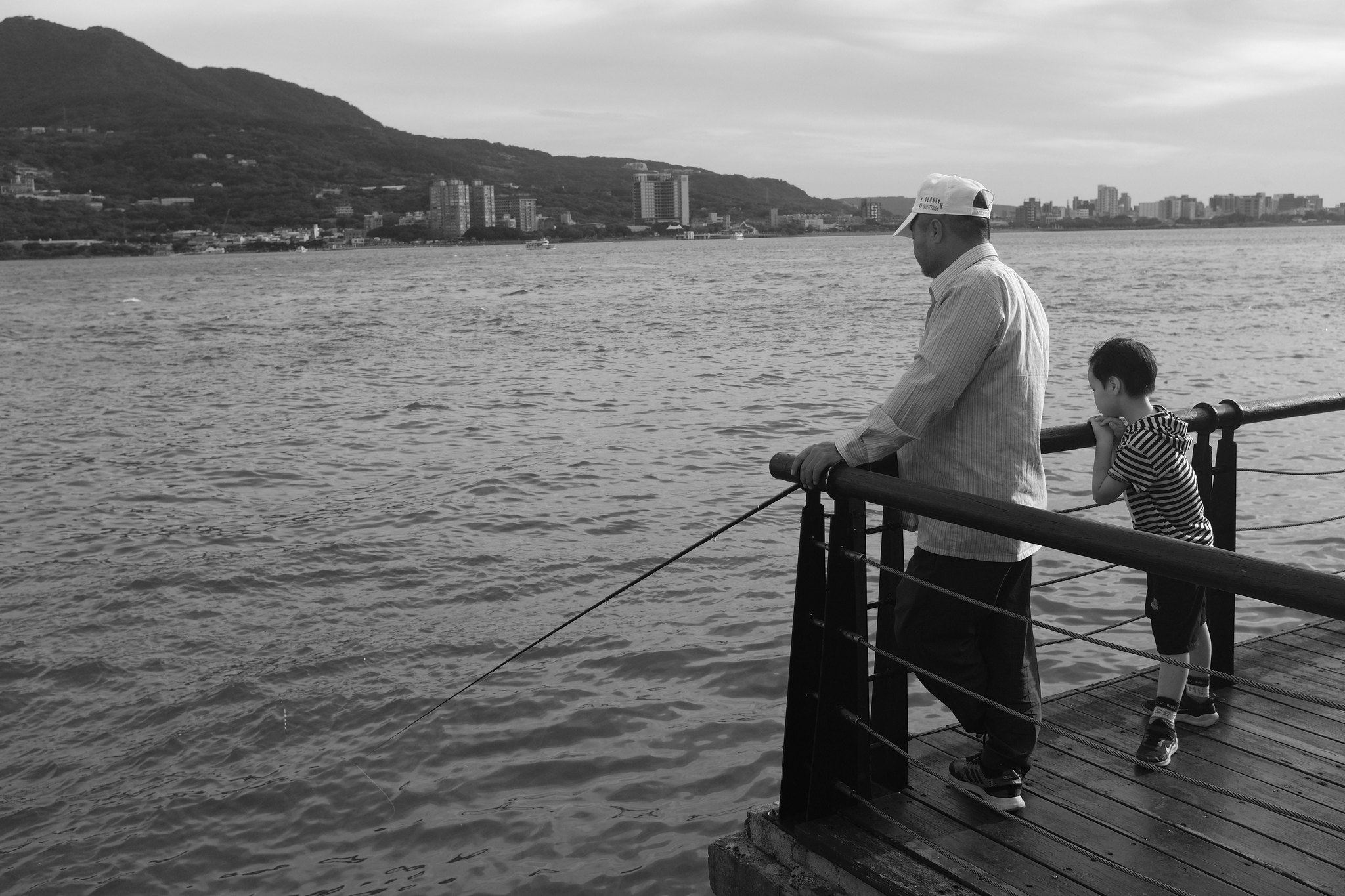 Black and white street photograph of an adult and child looking across the Tamsui waterfront from a wooden pier
