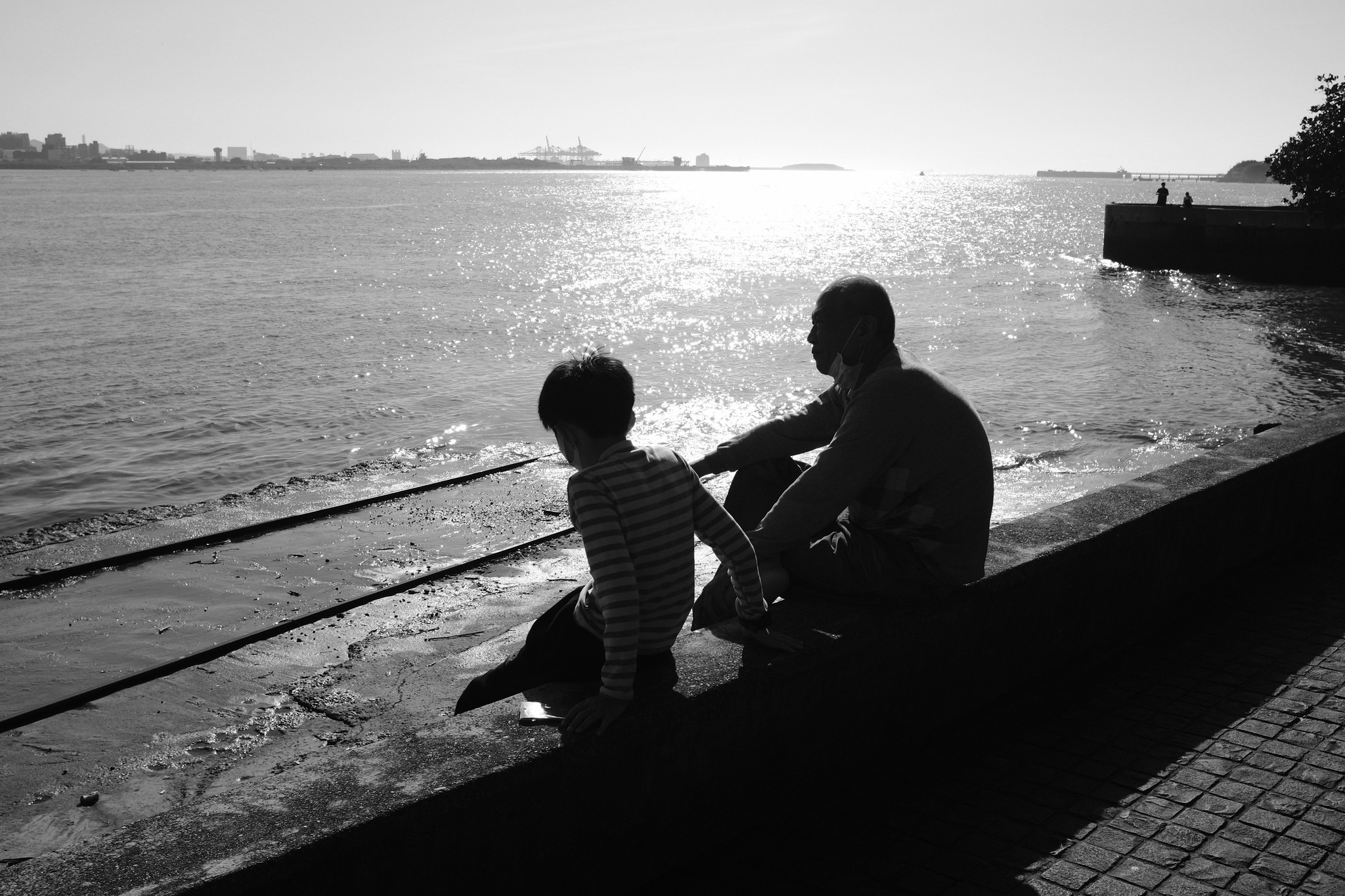 Black and white waterfront scene of a child and elder sitting beside the bright river in Tamsui, Taipei