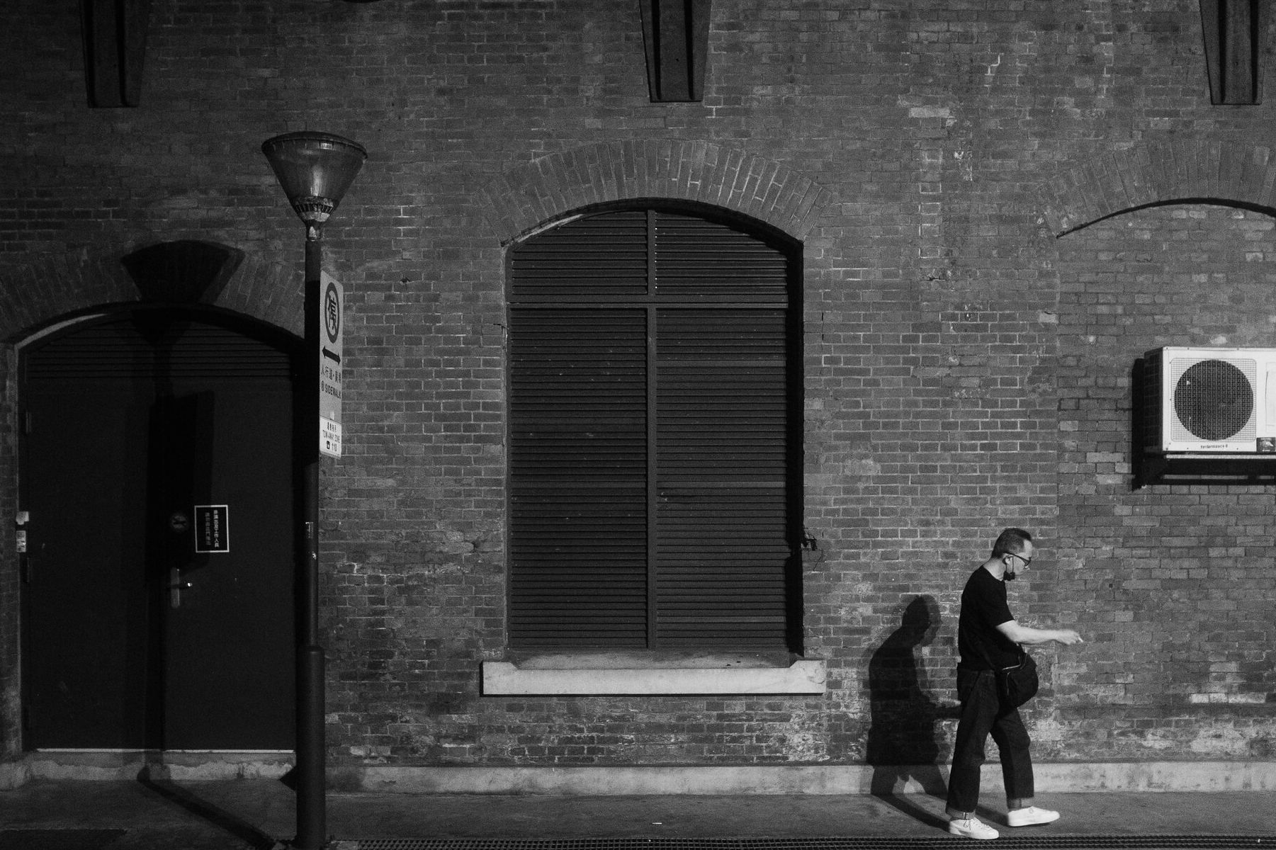 Black and white street photograph of a pedestrian walking beside a shuttered window on a brick wall in Ximending, Taipei