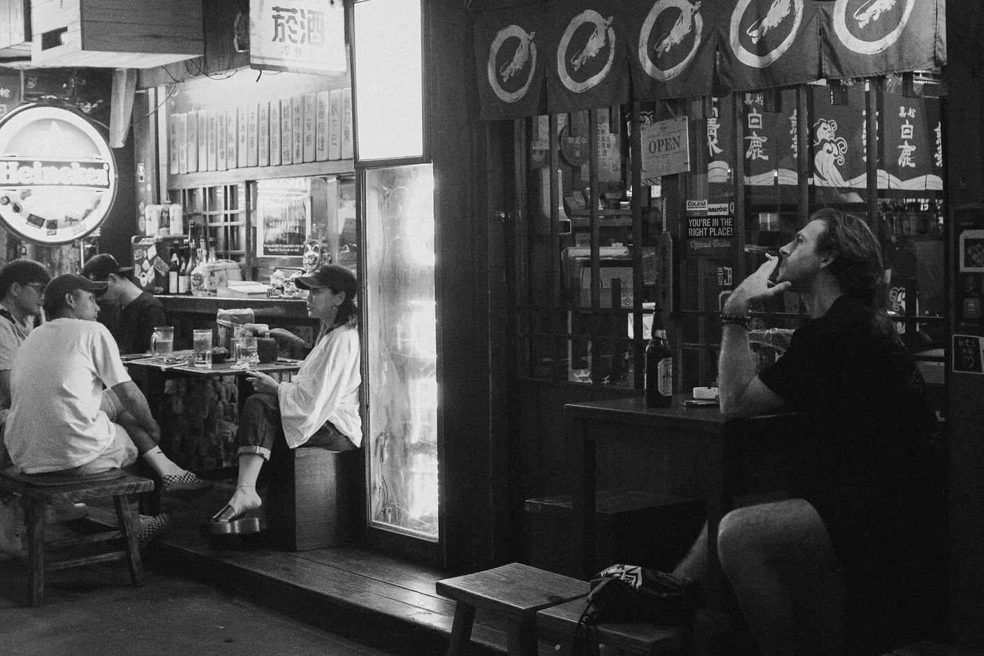 Black and white candid night scene outside a Ximending bar, with friends at tables and a man smoking beside the storefront
