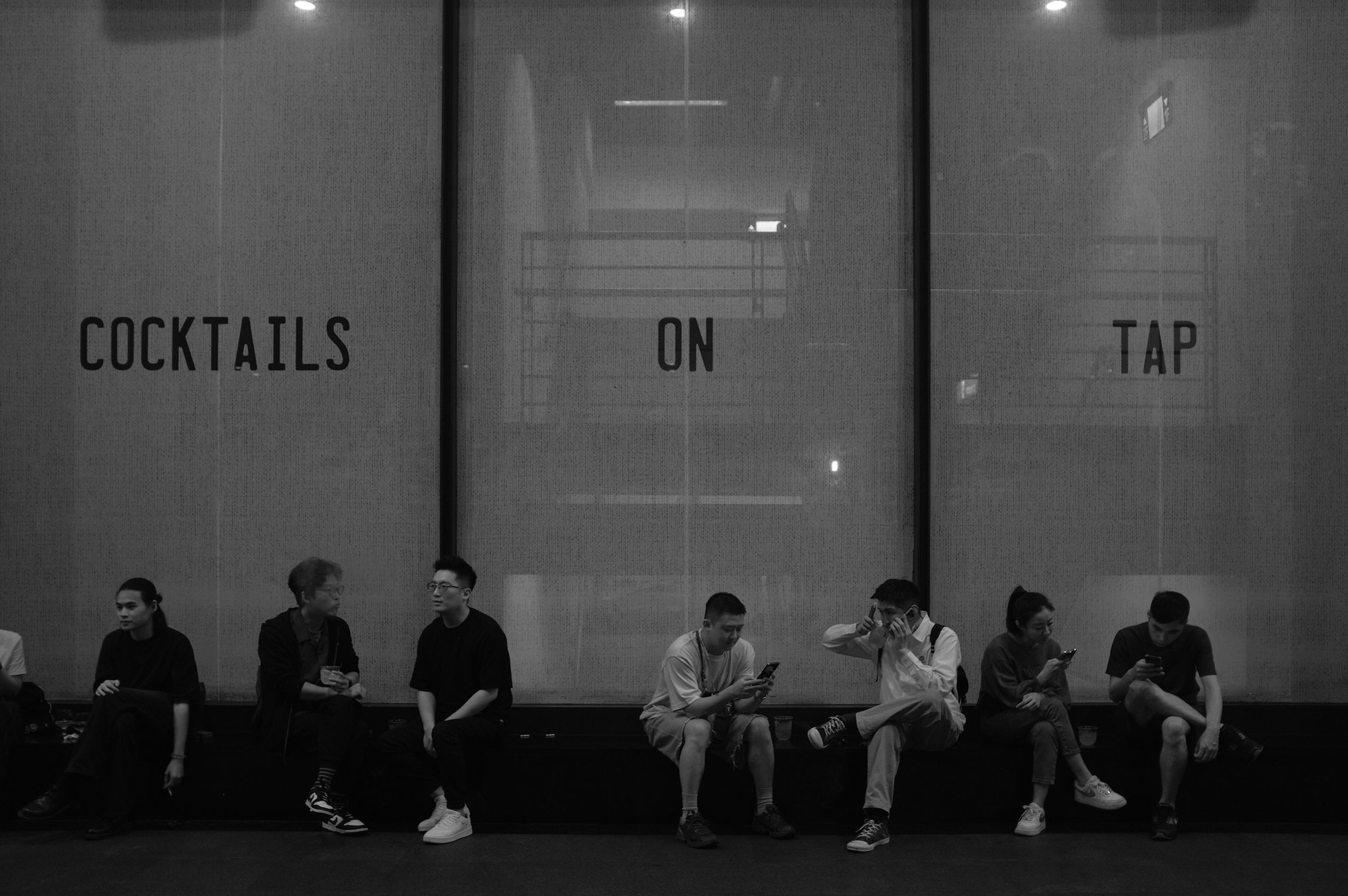 Black and white street photo of people sitting beneath a Cocktails on Tap window sign in Xinyi, Taipei