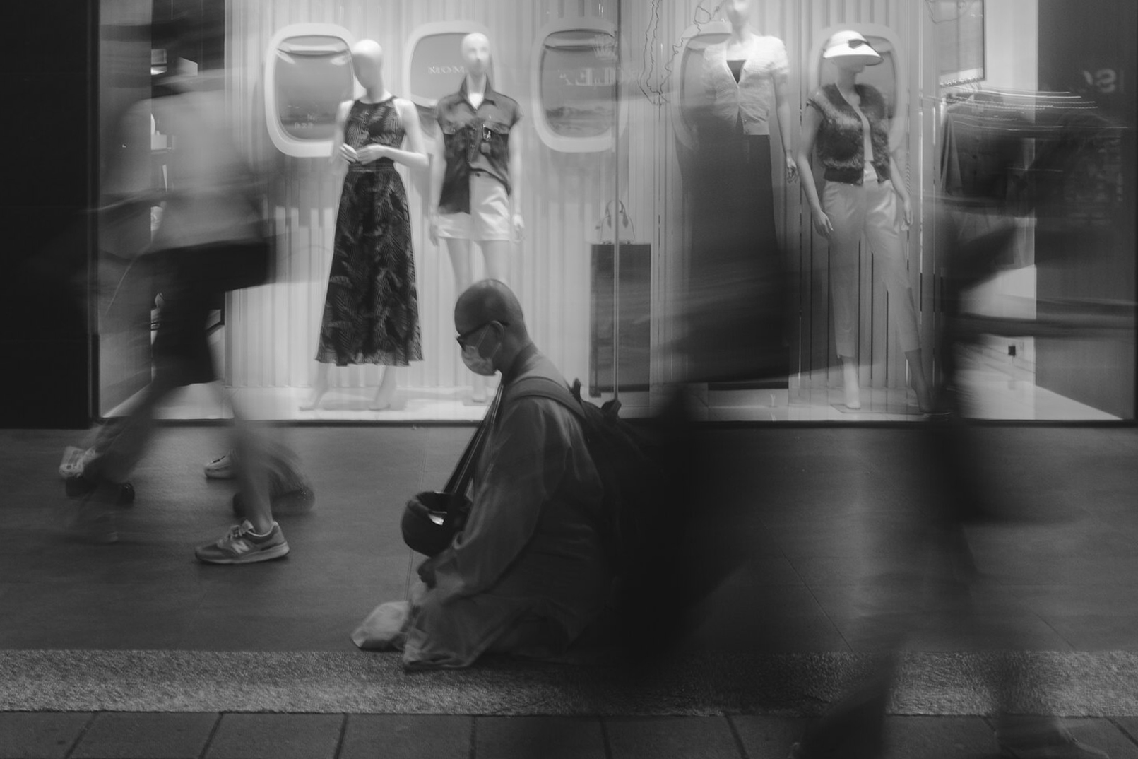 Black and white Zhongshan, Taipei street scene with a masked man seated in front of a boutique window display while blurred pedestrians pass by